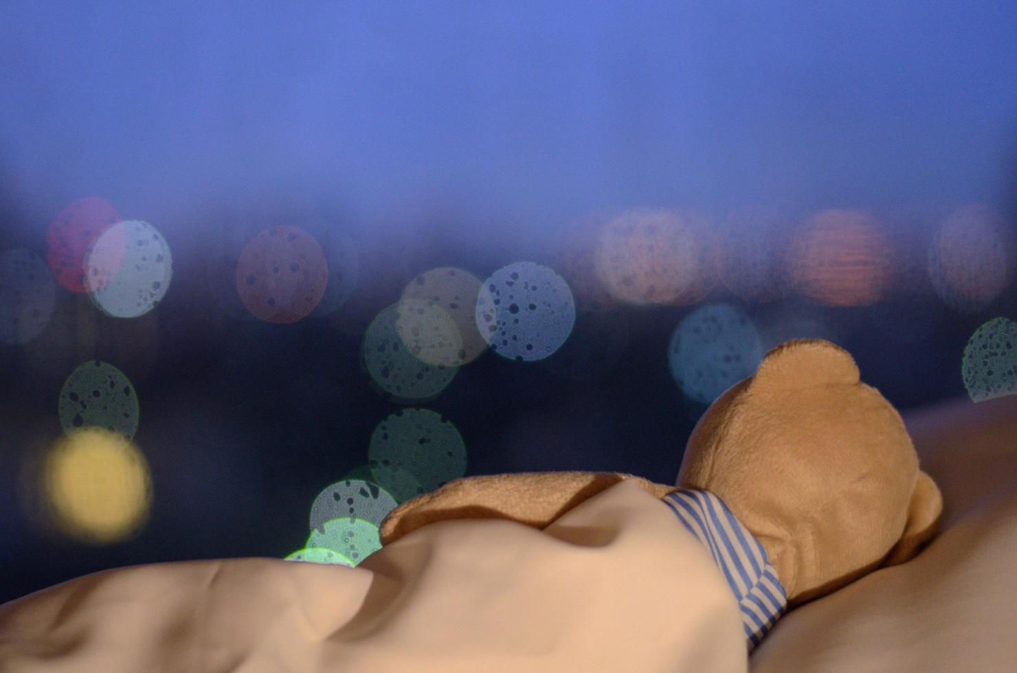 ursinho dormindo sozinho na cama com travesseiro branco e cobertor de frente para a janela com gota de chuva e luz bokeh na noite solitária. foto