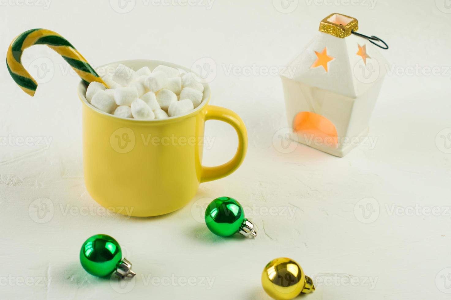 caneca amarela com chocolate quente de natal e marshmallow, com caramelo em um fundo branco com bolas de vidro e uma vela acesa. foto