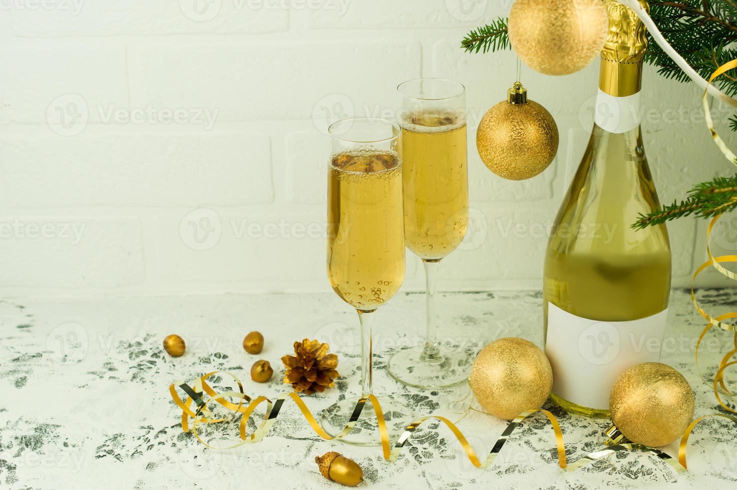 uma garrafa de champanhe e duas taças de vinho espumante na árvore de natal com bolas douradas em uma mesa branca por uma parede de tijolos. foto