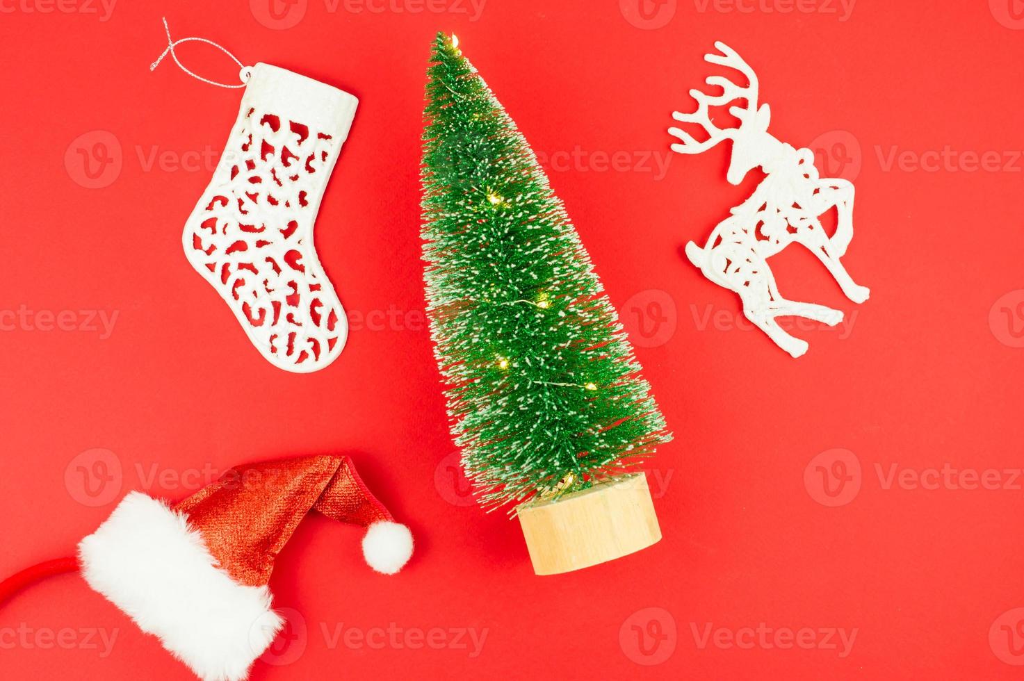 disposição plana. vista do topo. sobre um fundo vermelho, uma árvore de brinquedo com luzes, um chapéu de Papai Noel, um veado branco e uma meia de natal. foto