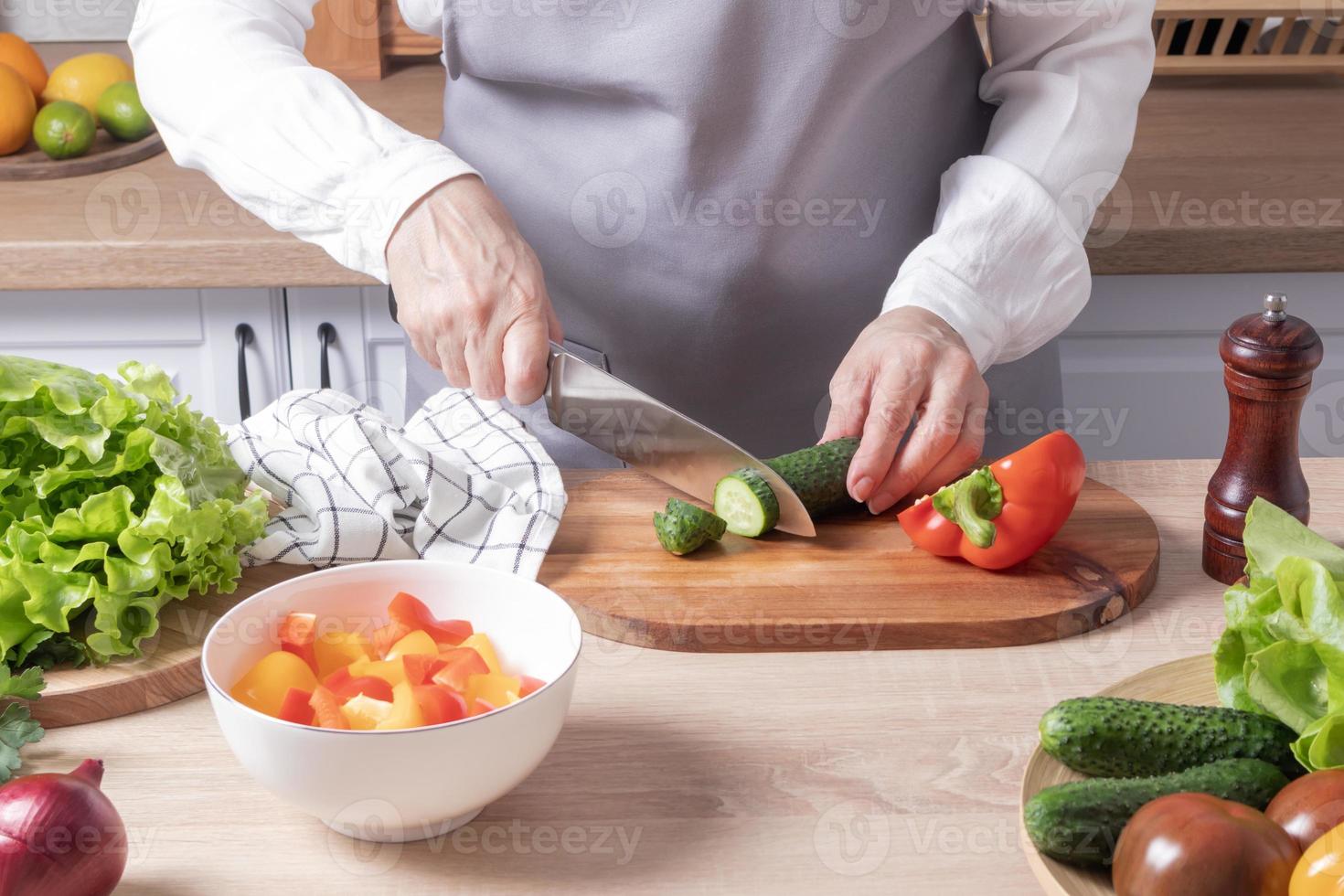 uma mulher idosa em um avental cinza corta um pepino verde fresco em uma tábua de corte em uma cozinha moderna. preparação de salada de legumes. foto