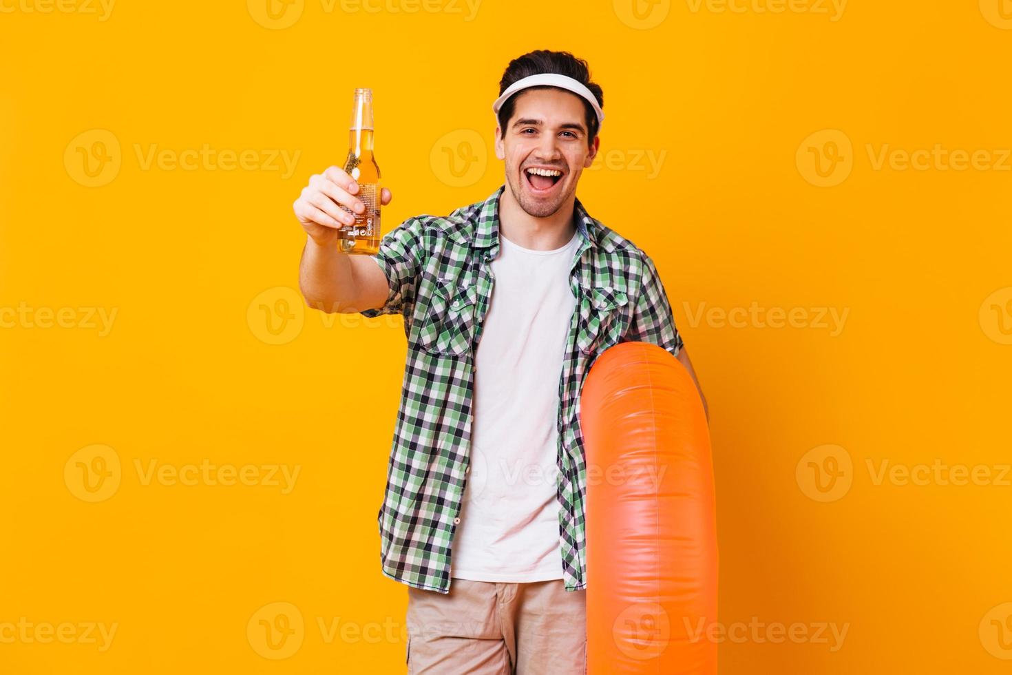 homem positivo com roupa de verão sorrindo e posando com garrafa de cerveja e círculo inflável foto
