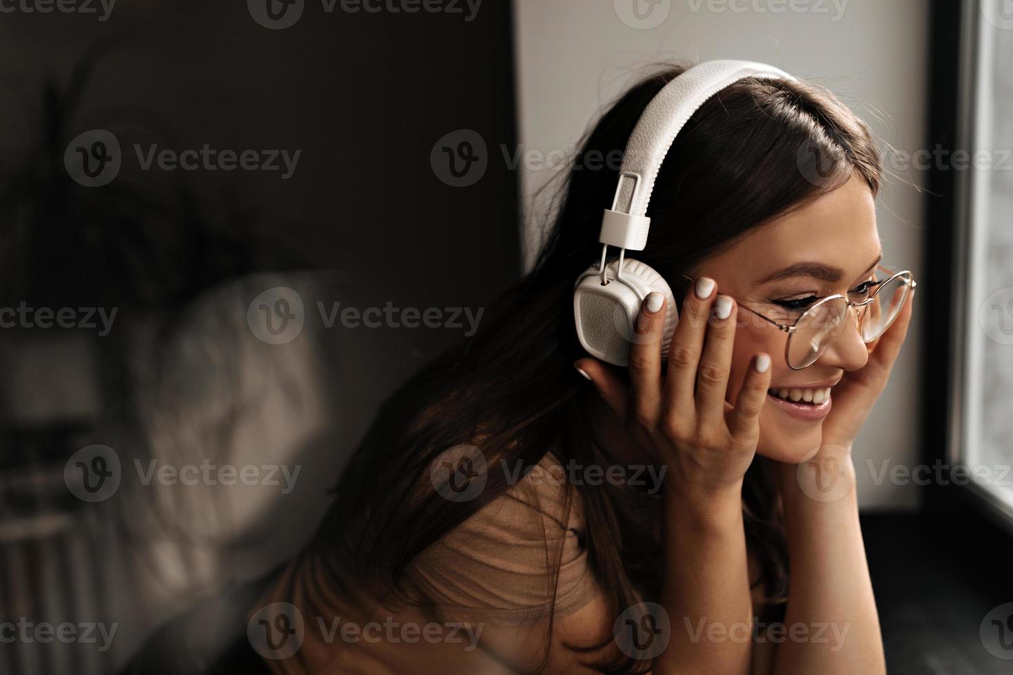mulher positiva em fones de ouvido maciços brancos coloca óculos e sorri, apoiando-se no peitoril da janela preta foto