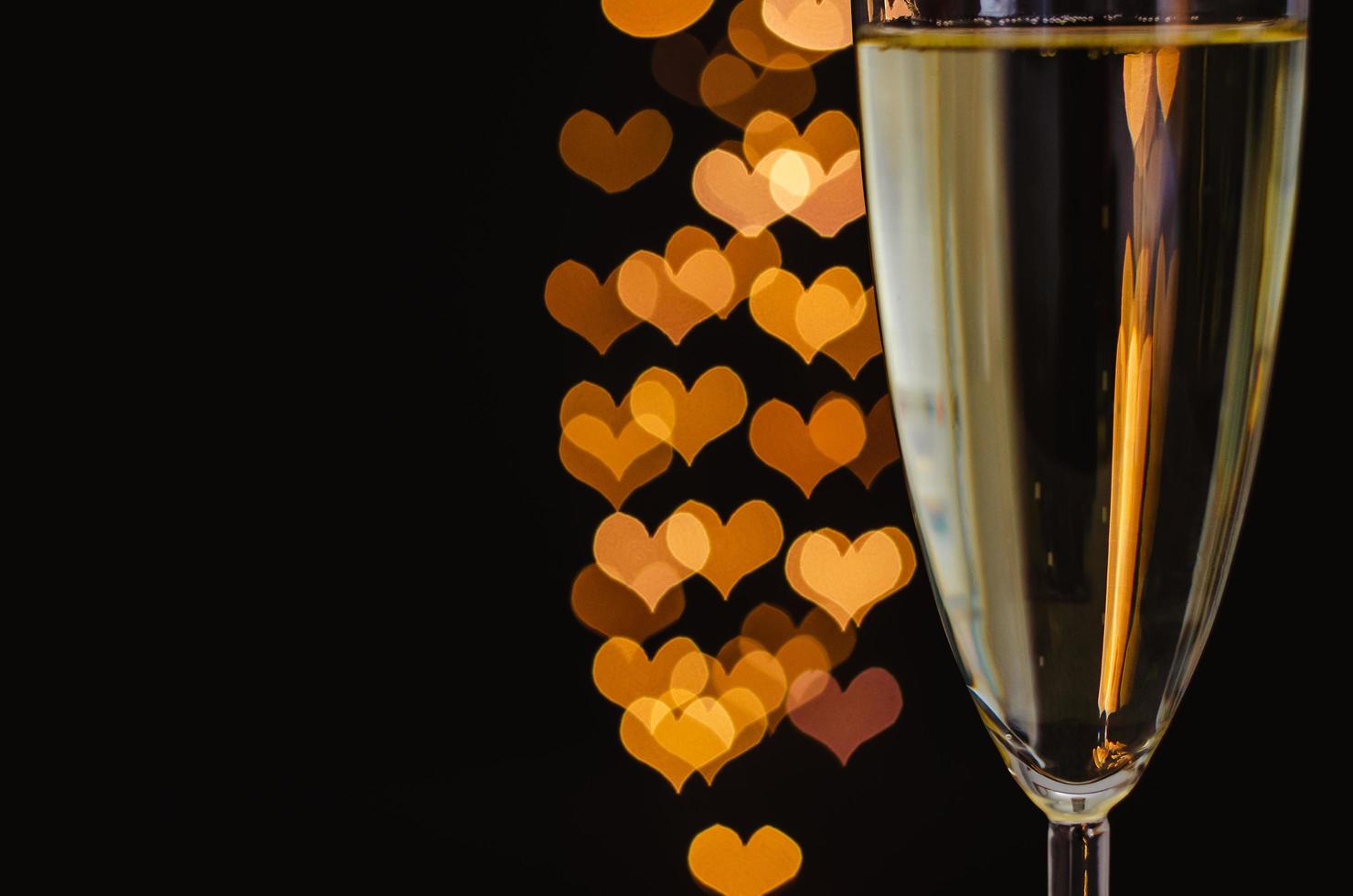 foco parcial de um copo de vinho espumante com luzes de bokeh de forma de amor dourado em fundo escuro. foto