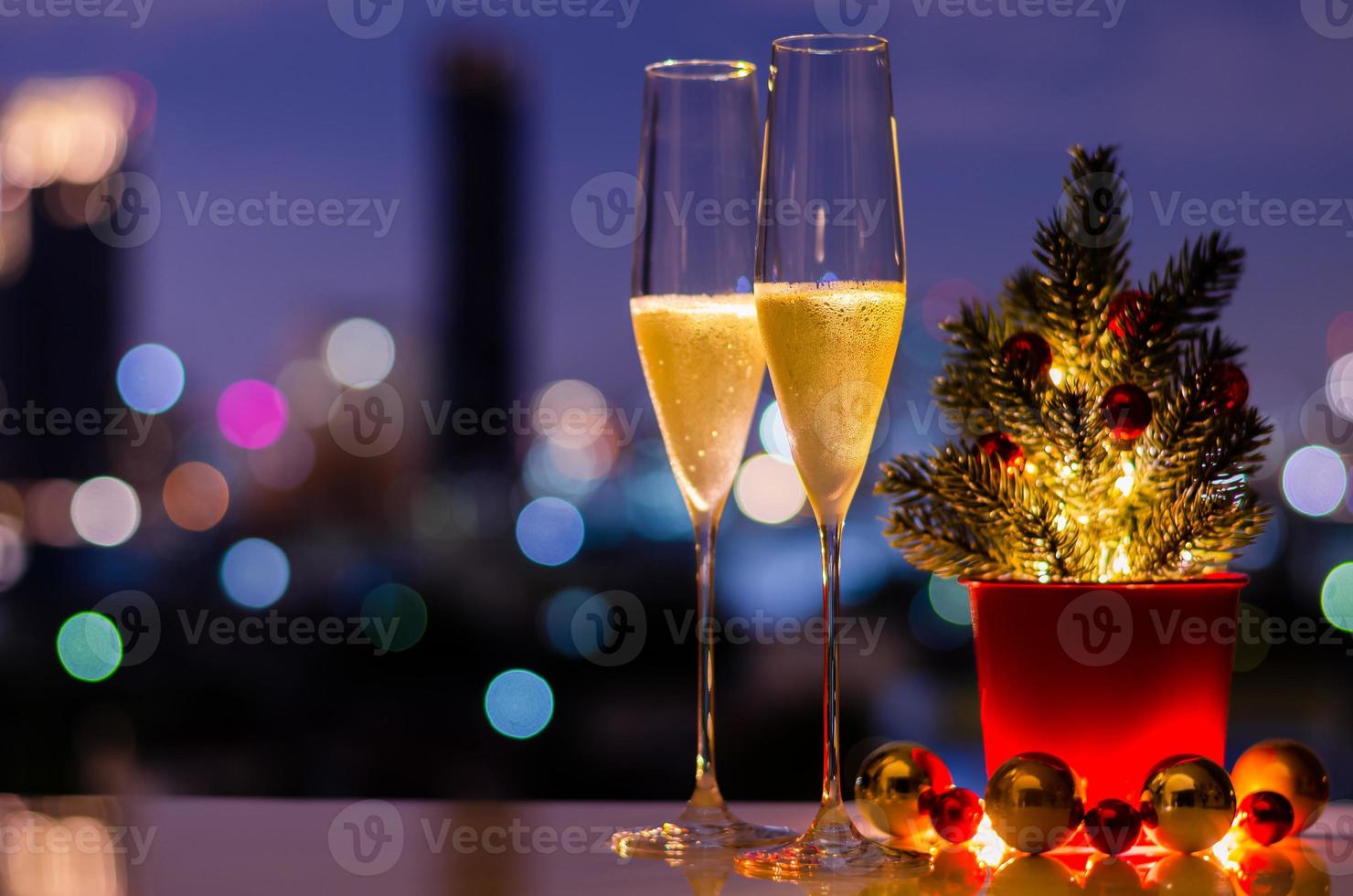 dois copos de vinho espumante com árvore de natal e enfeites de bugiganga colocados na mesa com fundo colorido de luzes de bokeh da cidade. foto