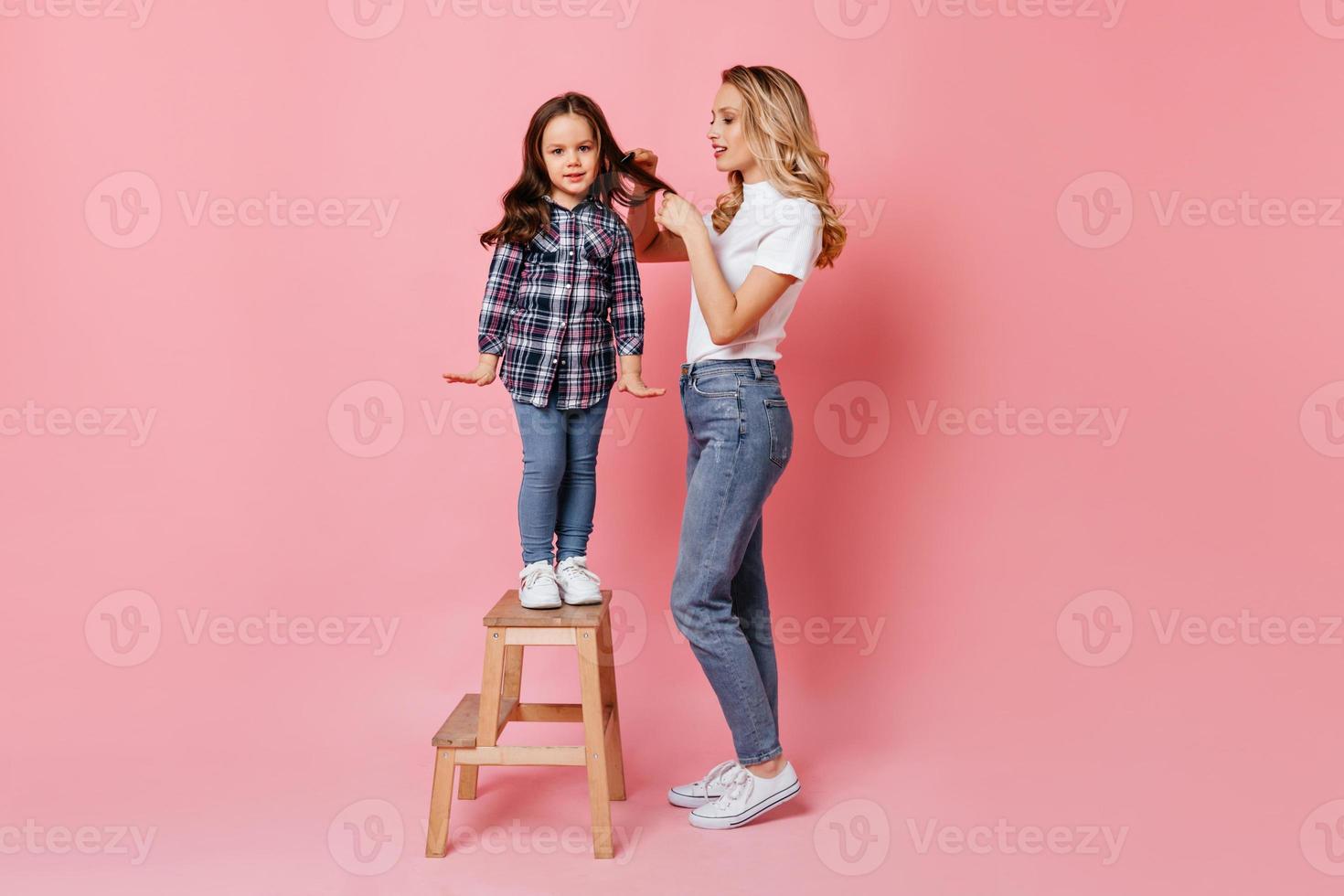 menina bonitinha na camisa xadrez está de pé na escada de madeira enquanto a mãe está escovando o cabelo foto