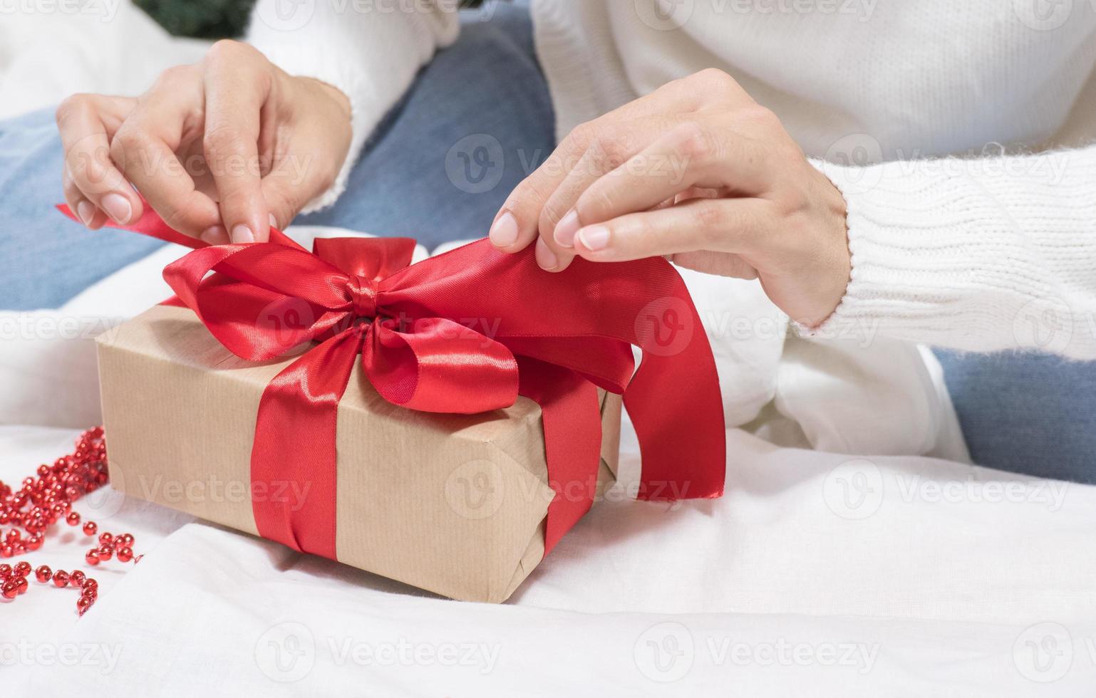 mãos femininas desamarram um laço em uma caixa de presente para o natal em uma cama branca. foto