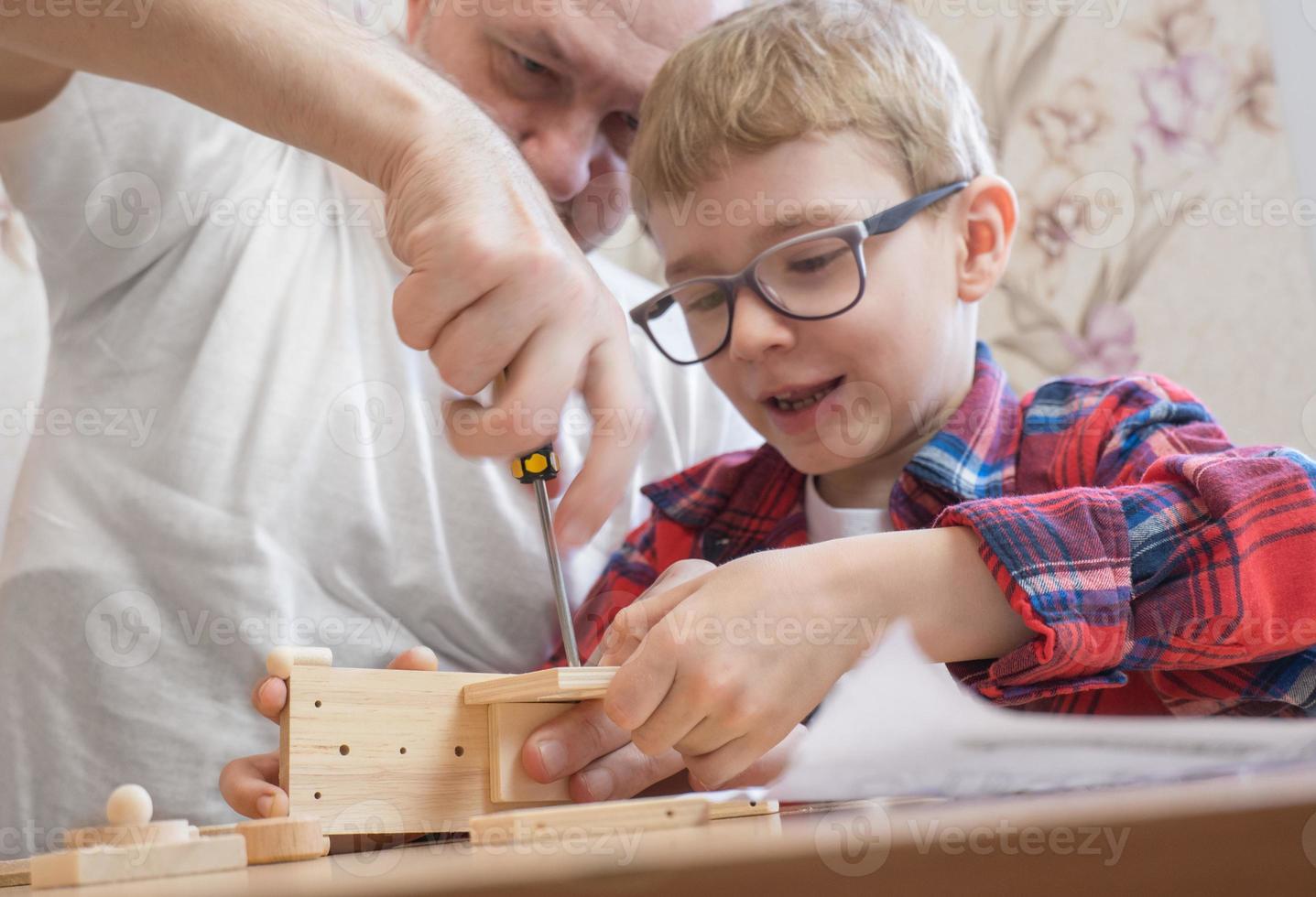 feliz dia dos pais e conceito de infância. pai e filho de óculos trabalham com ferramentas manuais, usando uma chave de fenda, montando um construtor de madeira em casa à mesa. foto