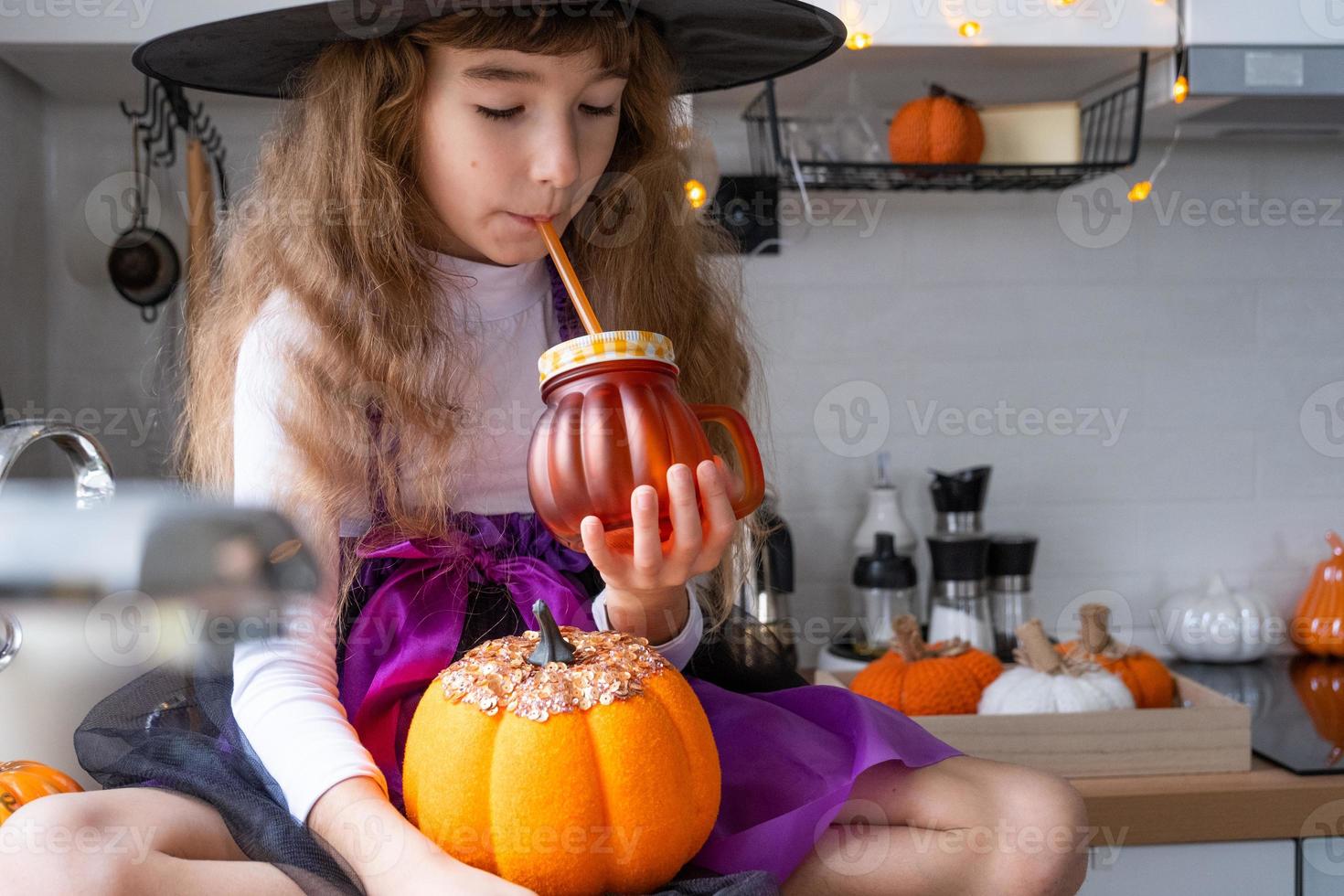 criança bebe suco de abóbora, smoothie de vegetais ou coquetel de uma caneca com um canudo. cozinha decora para o halloween. garota em uma fantasia de bruxa. outono aconchegante em casa foto