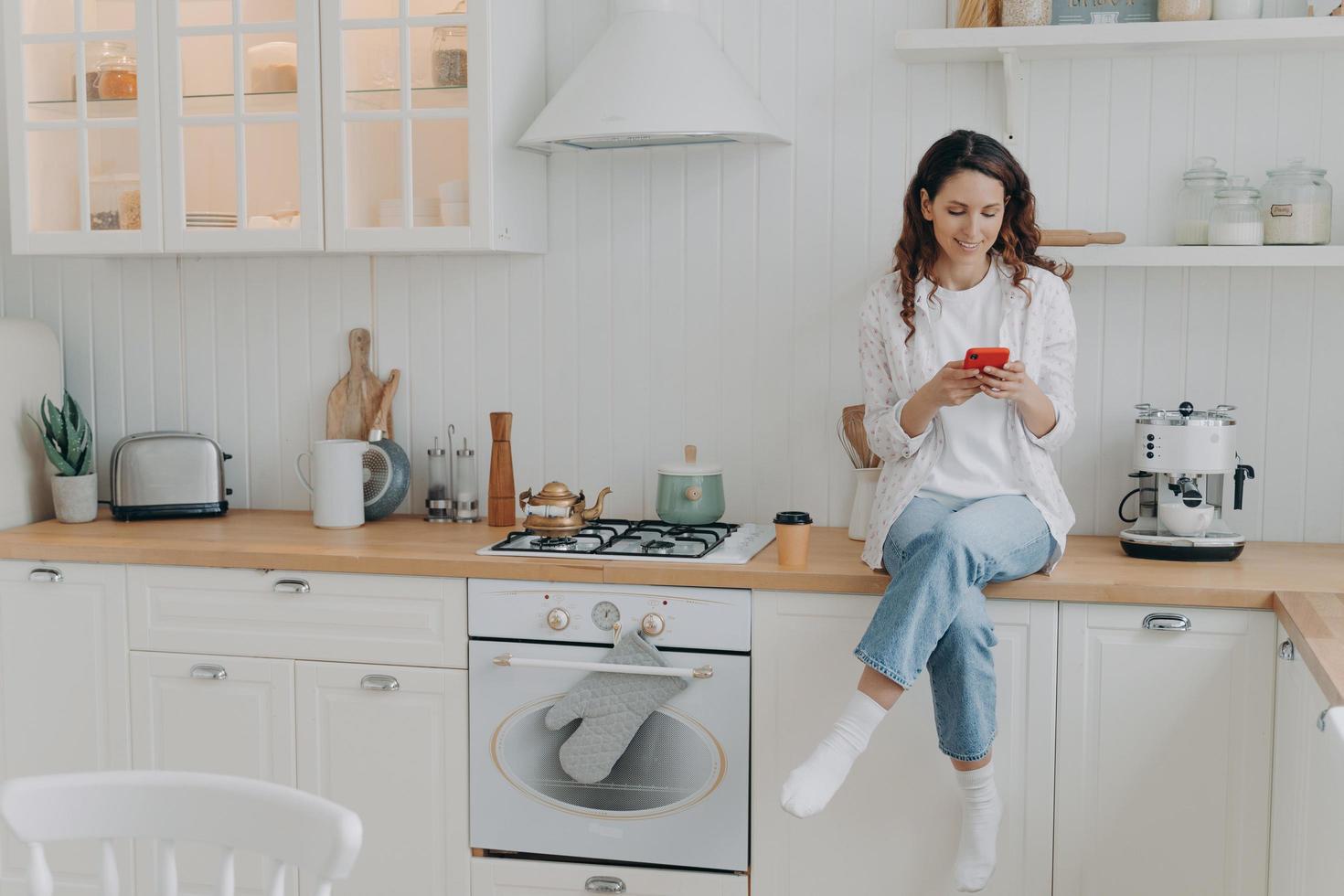dona de casa feminina satisfeita com os serviços de limpeza da casa senta-se na cozinha moderna e limpa com smartphone foto