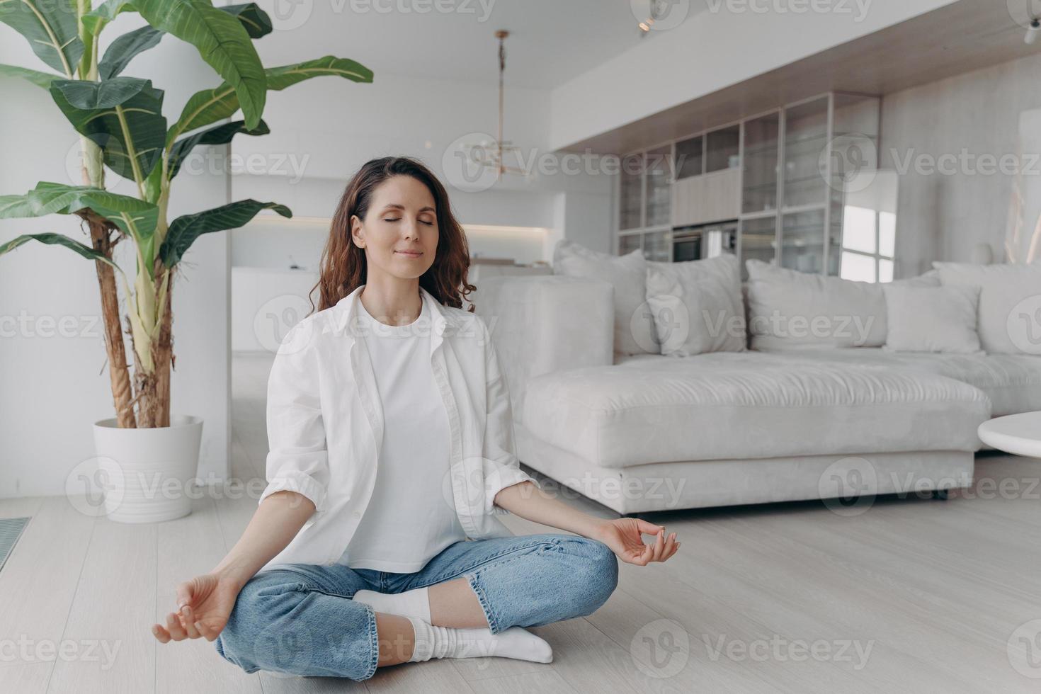 serena mulher praticando ioga sentada em postura de lótus no chão em casa. bem-estar, estilo de vida saudável foto