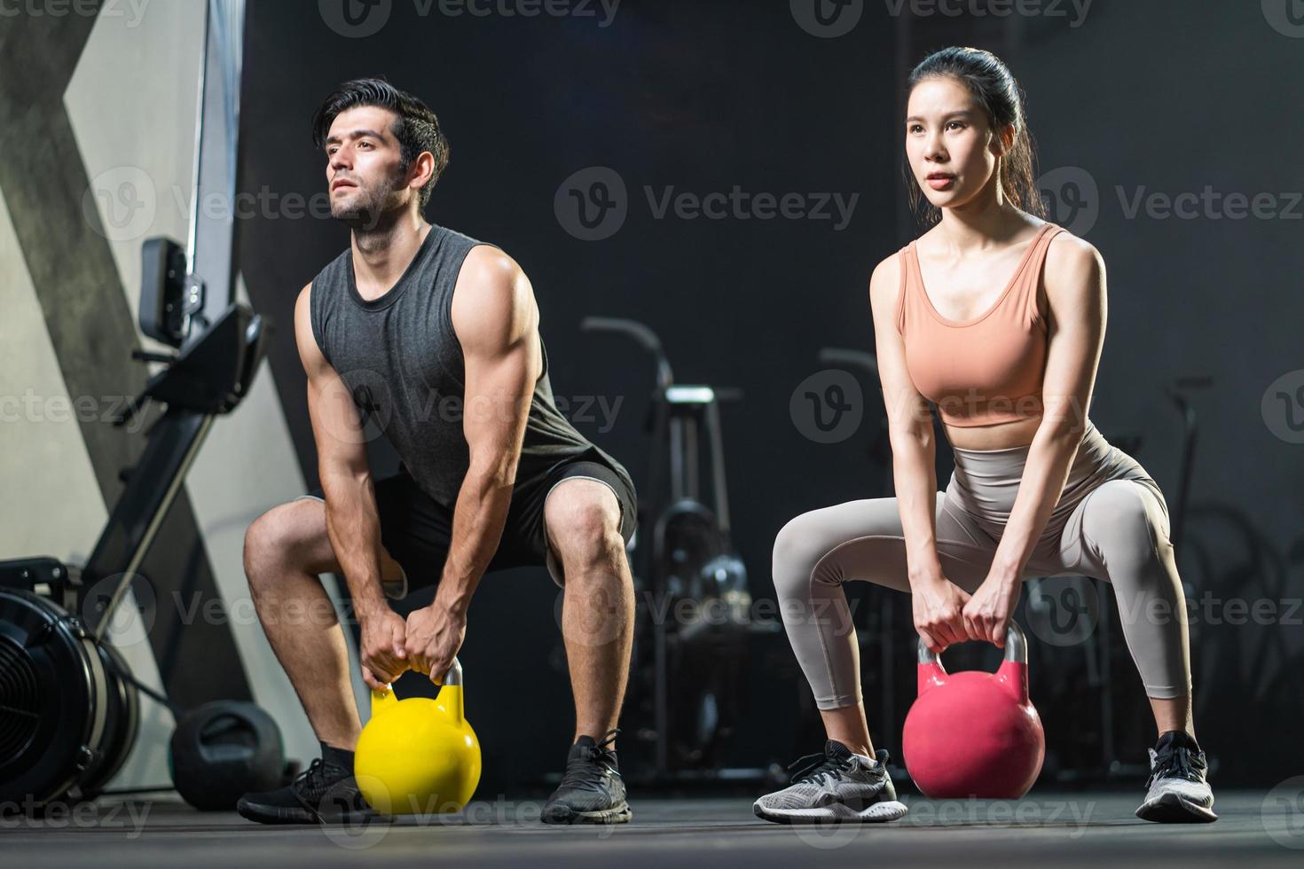 foco seletivo em mulheres e homens asiáticos ao fundo. os homens balançam o kettlebell para fazer exercícios de crossfit e levantamento de peso dentro da academia de ginástica para treinar a força do corpo físico e os músculos dos braços firmes foto