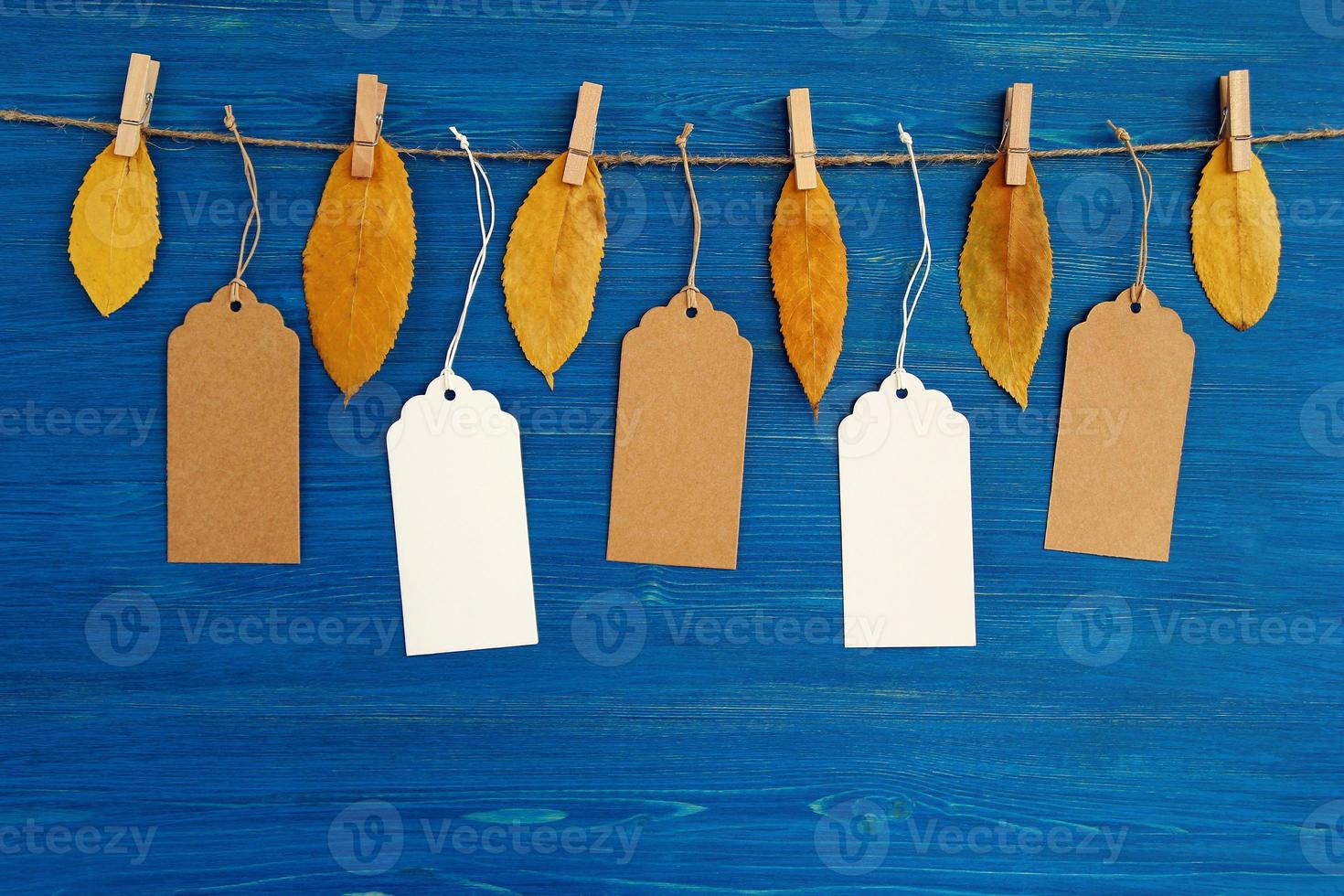 cinco etiquetas de preço ou etiquetas de papel em branco marrom e branco penduradas em uma corda com folhas de outono amarelas secas no fundo azul de madeira. foto