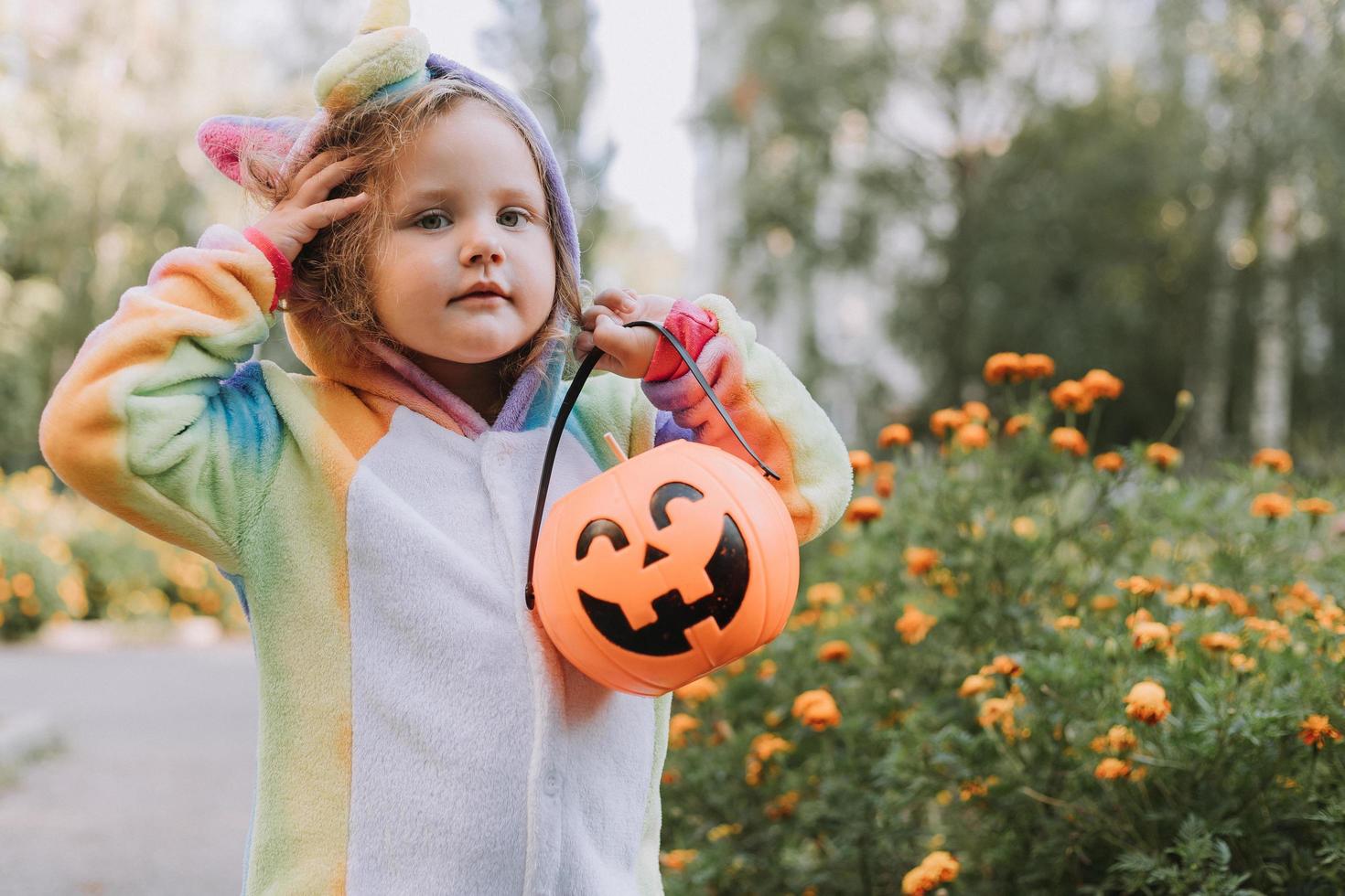 menina bonitinha em uma fantasia de unicórnio arco-íris para o halloween vai coletar doces em uma cesta de abóbora em uma área residencial. criança caminha ao ar livre. doçura ou travessura. estilo de vida. kigurumi foto