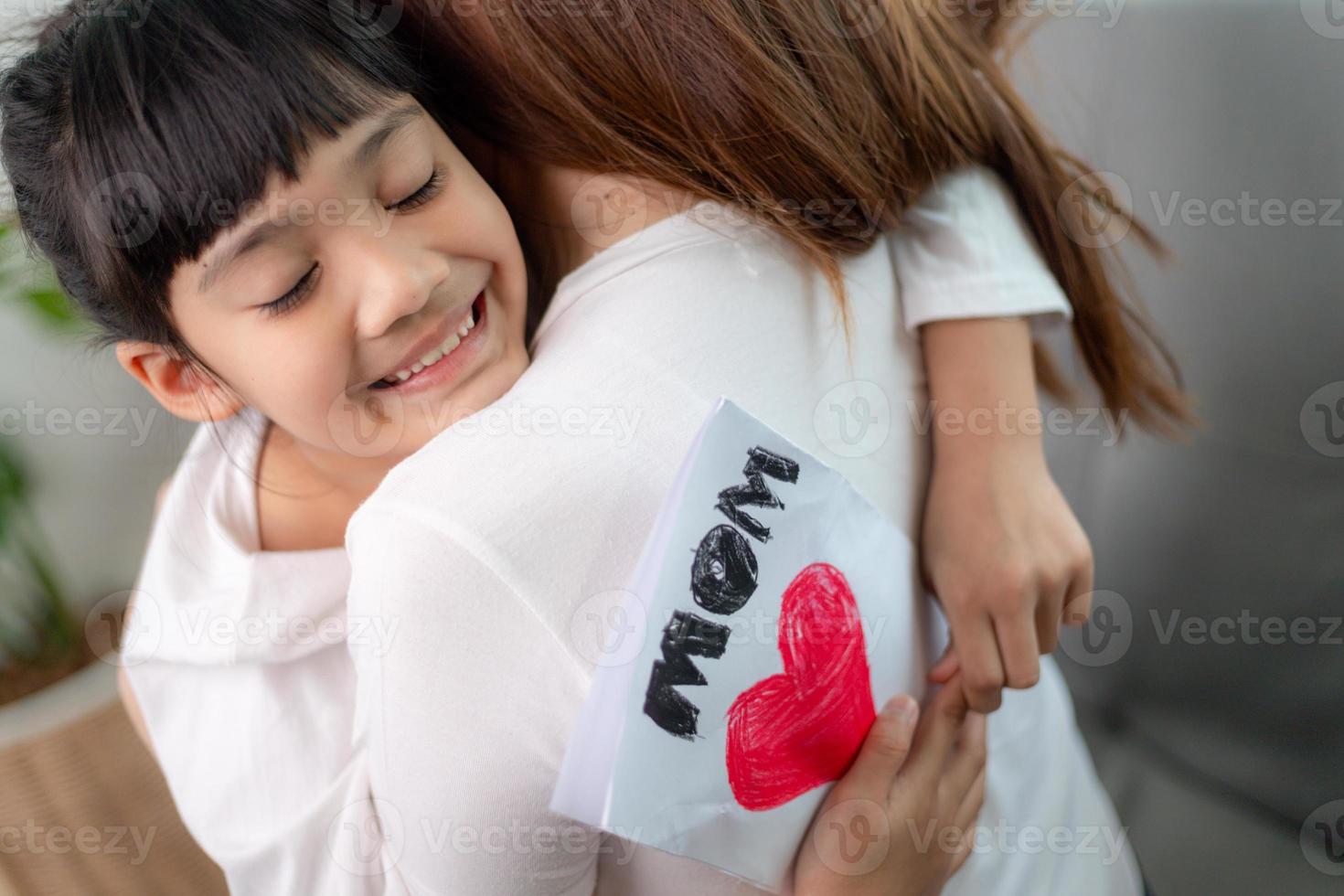 A adorável filha pré-escolar doce asiática parabeniza a mamãe com o feriado de aniversário do evento de vida, prepara-se para seu cartão postal artesanal com símbolo de coração pintado de vermelho de amor incondicional, conceito de dia das mães foto