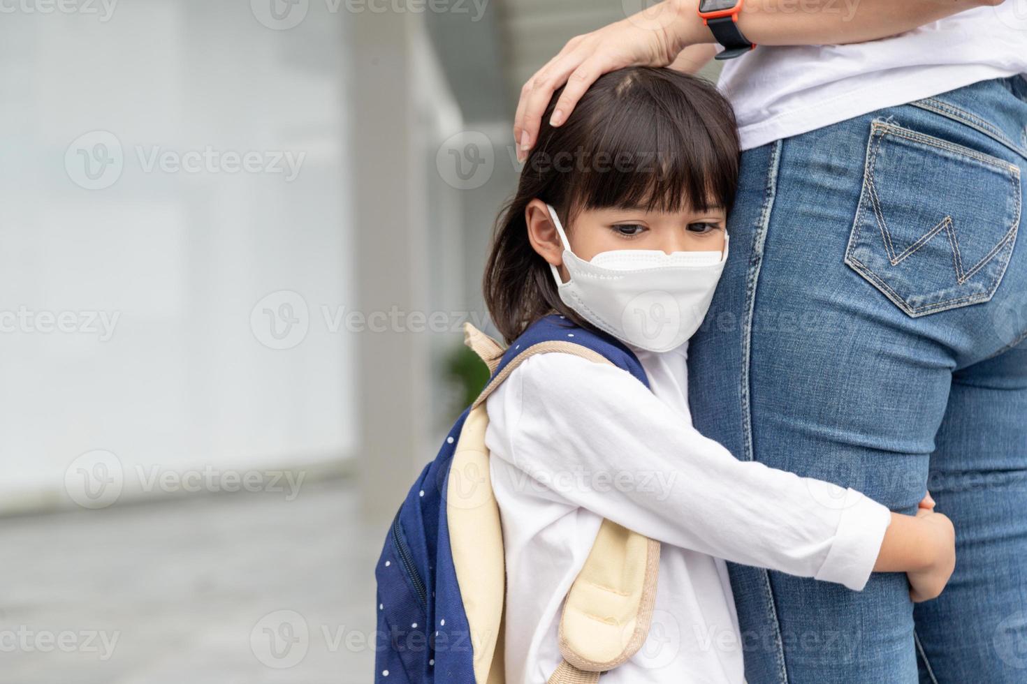 mãe acompanha a criança à escola. a mãe apoia e motiva o aluno. a garotinha usando uma máscara facial não quer deixar a mãe. teme a escola primária. foto