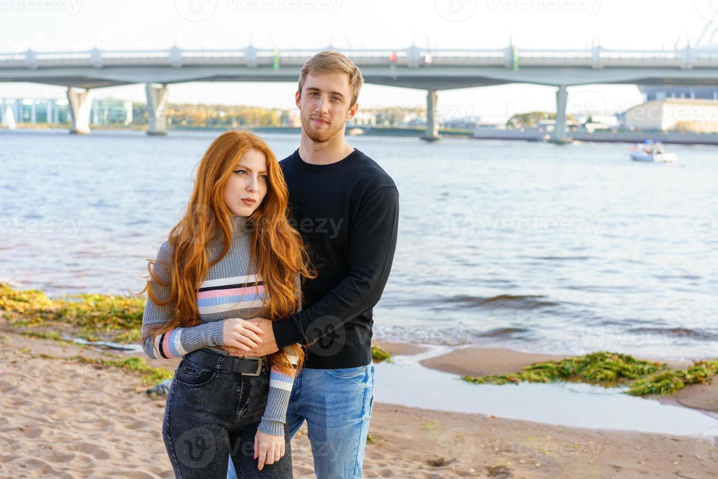 jovem casal está de pé na margem do rio abraçando foto