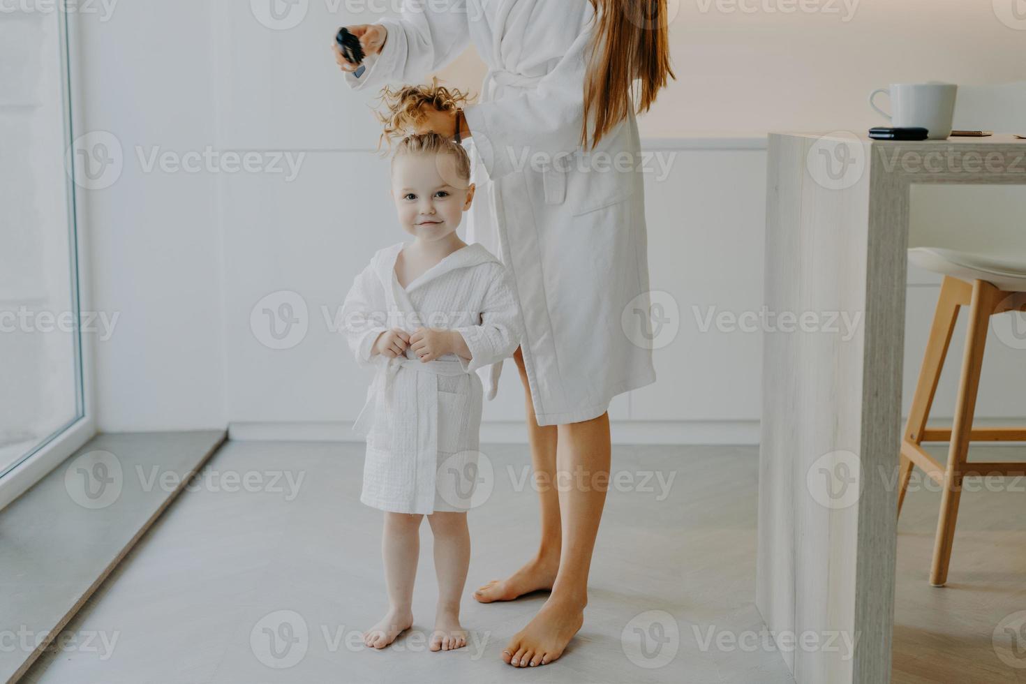 mãe sem rosto faz rabo de cavalo do cabelo encaracolado da filha usa pente. menina de três anos bonitinha em roupão branco depois de tomar banho posa perto da mãe em casa. tempo de beleza da paternidade das crianças foto