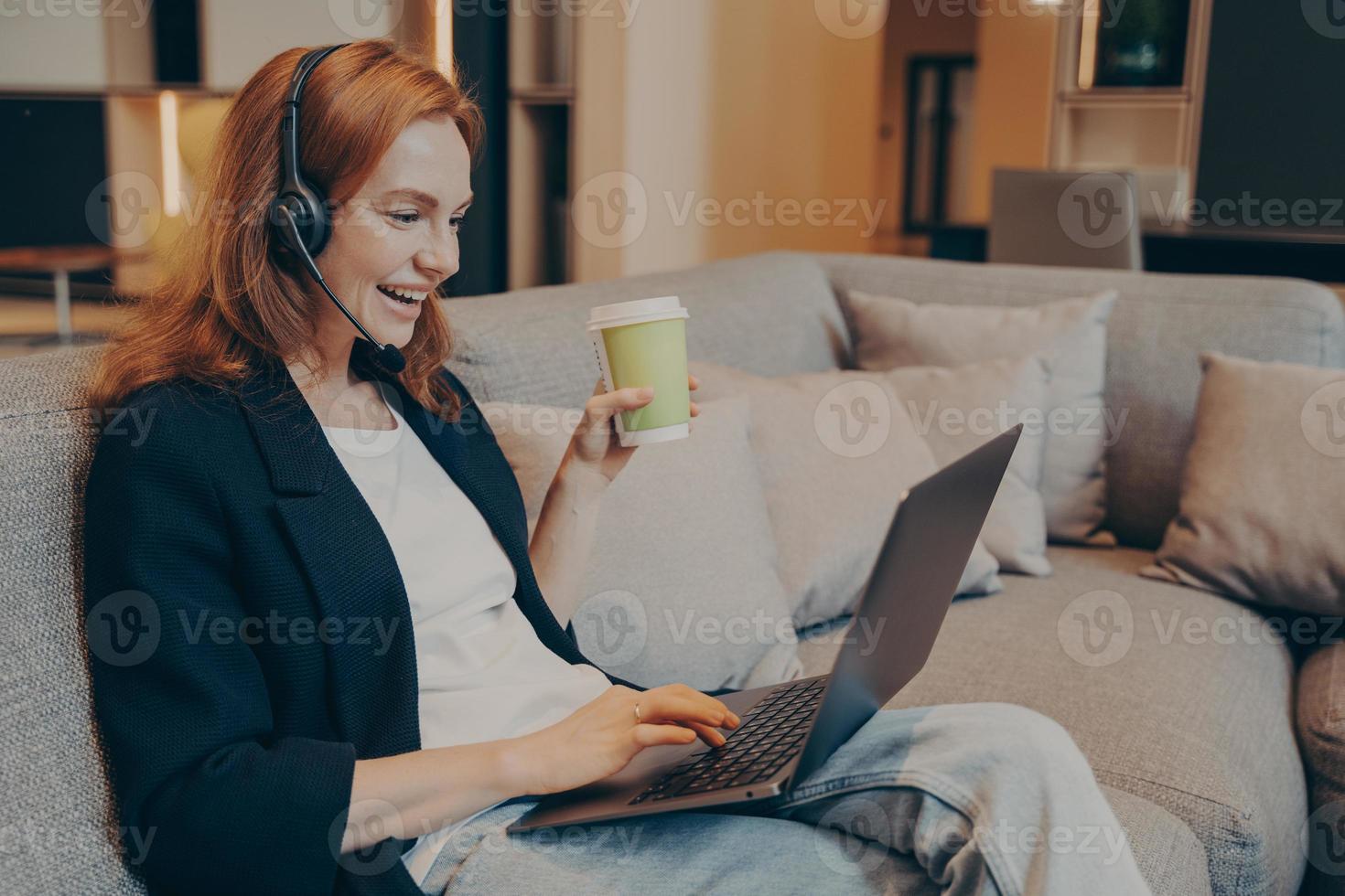 mulher sorridente ruiva animada com fone de ouvido sem fio e laptop sentado no sofá no café foto