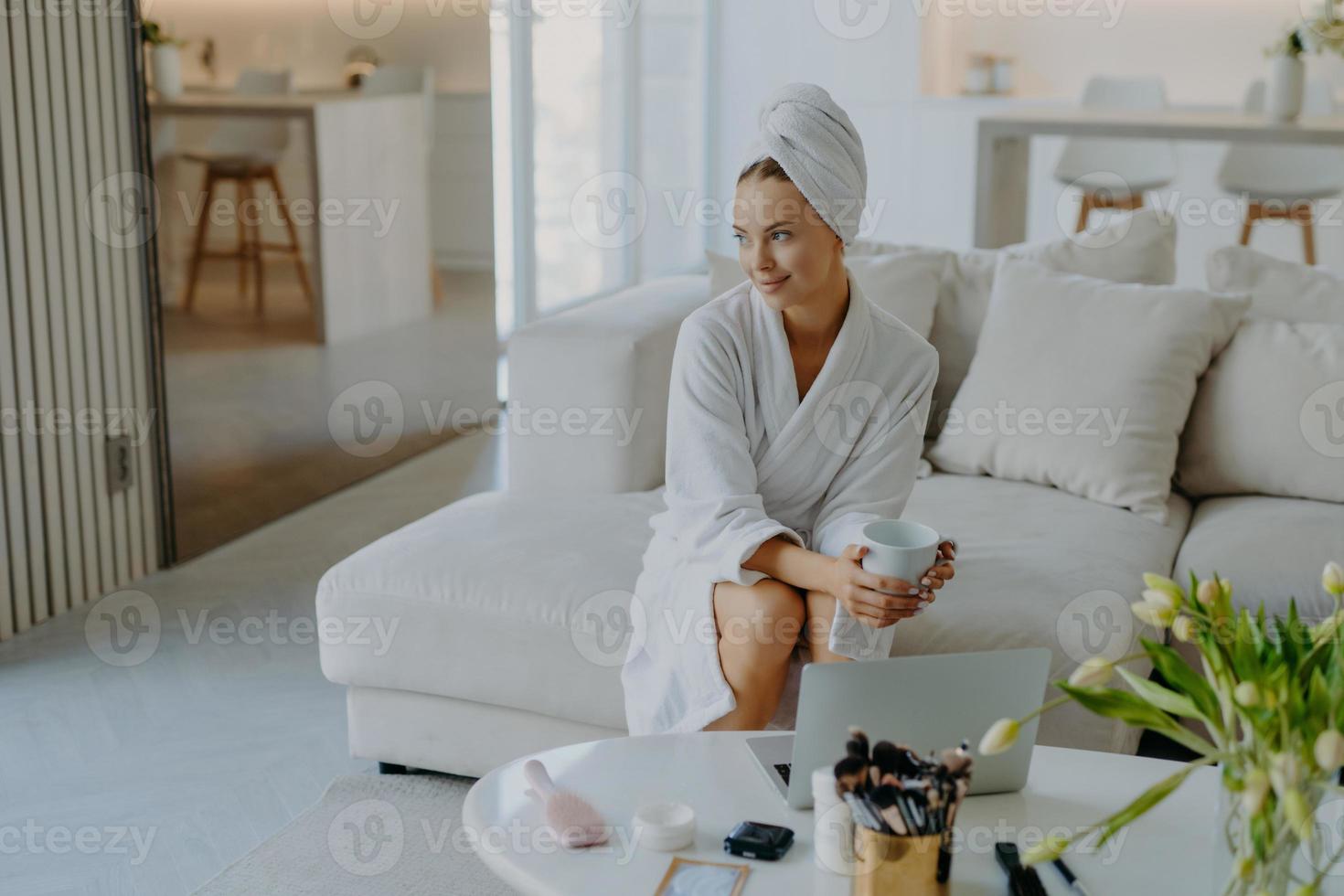 mulher pensativa relaxada, vestida de roupão de banho e toalha enrolada na cabeça, senta-se no sofá com uma xícara de bebida perto da mesa com produtos cosméticos parece pensativa ao lado de poses contra o interior aconchegante da casa foto