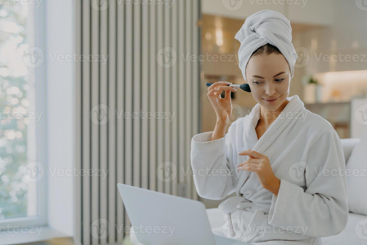 foto de jovem europeu aplica pó facial com escova cosmética concentrada no computador portátil faz videochamada tem o próprio blog de beleza posa em roupão confortável contra o interior da casa