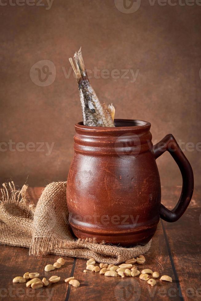 caneca marrom de barro de cerveja com cerveja e peixe seco em uma mesa de madeira em um fundo escuro. foto