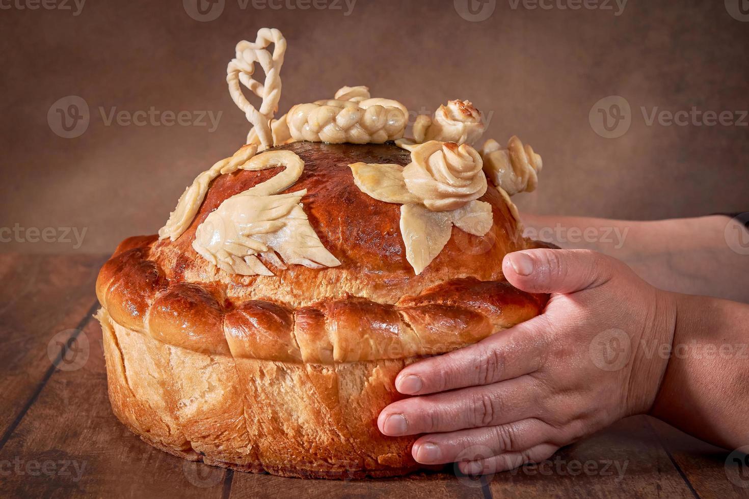 pão redondo de casamento russo em uma mesa de madeira é realizado pelas mãos de uma mulher gorda idosa. cerimônia de casamento russo. foto