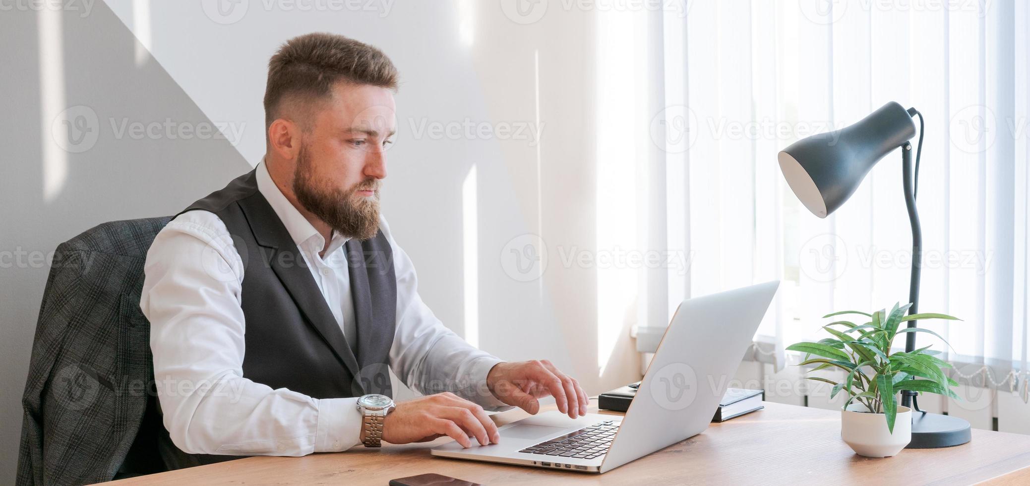 homem caucasiano senta-se à mesa perto de laptop no escritório e olha para a tela. sério foto