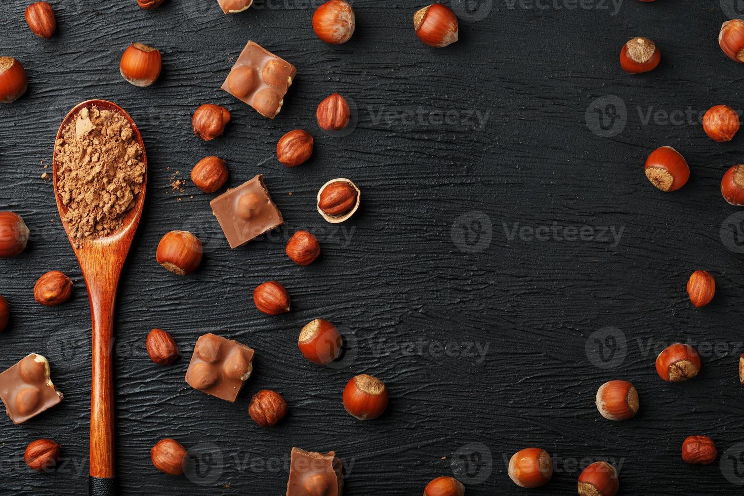 chocolate com avelãs e uma colher de pau com cacau em um fundo escuro, cercado por nozes com casca e descascadas. foto