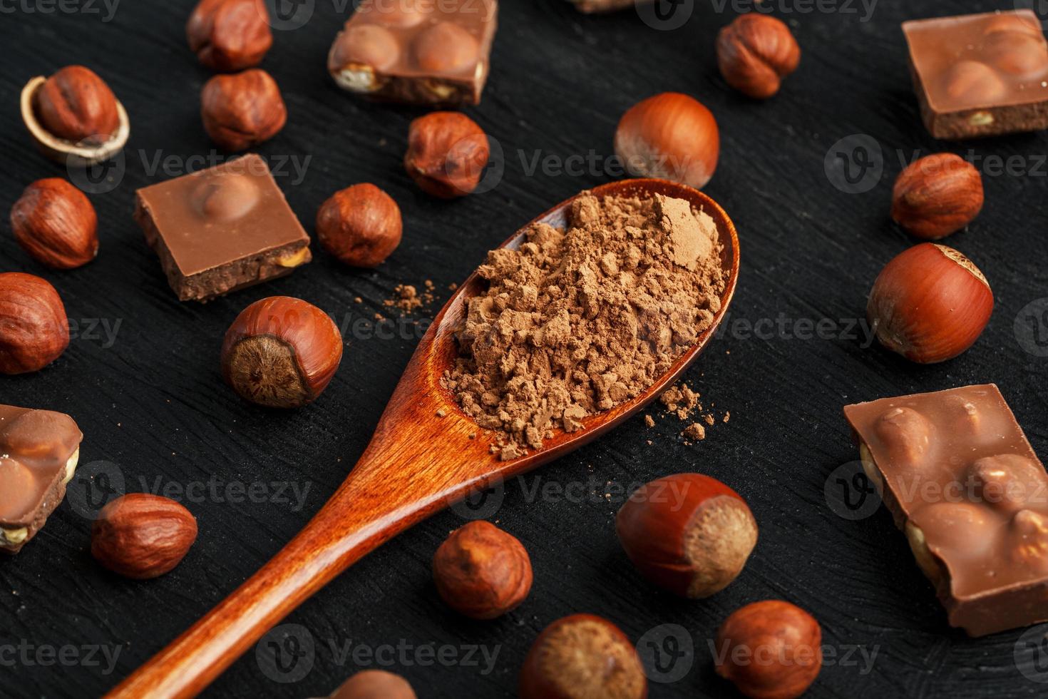 chocolate com avelãs e uma colher de pau com cacau em um fundo escuro, cercado por nozes com casca e descascadas. foto