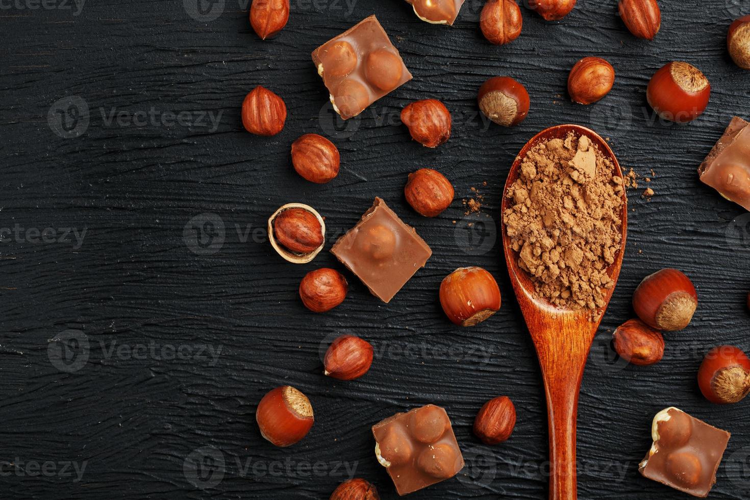 chocolate com avelãs e uma colher de pau com cacau em um fundo escuro, cercado por nozes com casca e descascadas. foto