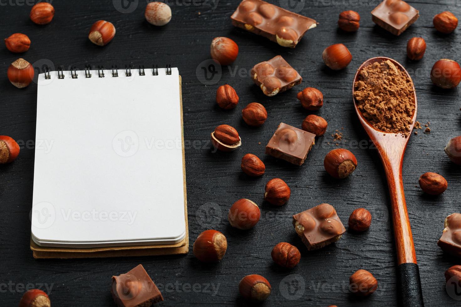 chocolate com avelãs, um caderno com páginas em branco e uma colher de pau com cacau em um fundo escuro, cercado por nozes com casca e descascadas. foto