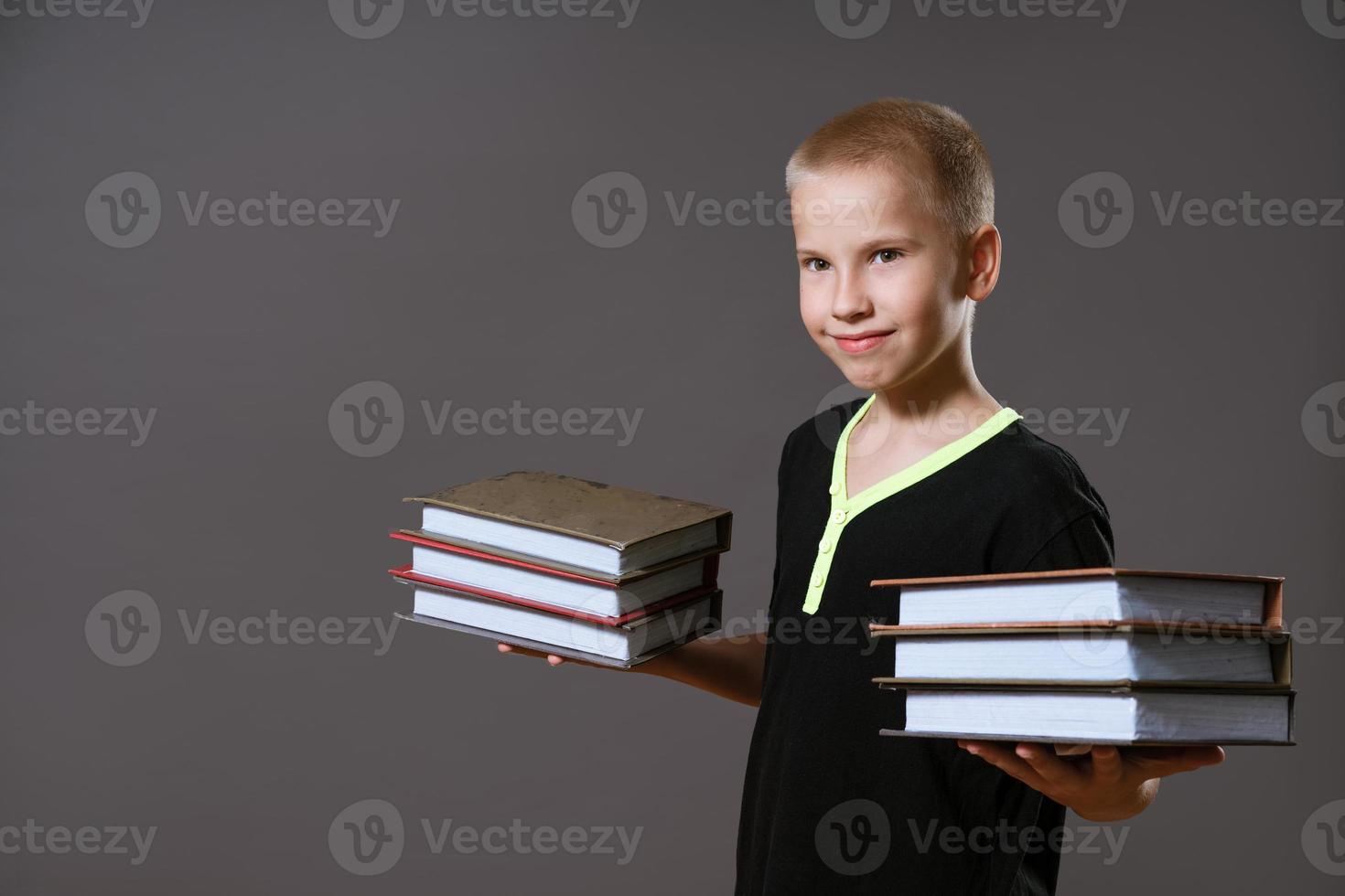 menino bonito segurando pilhas de livros em fundo cinza foto