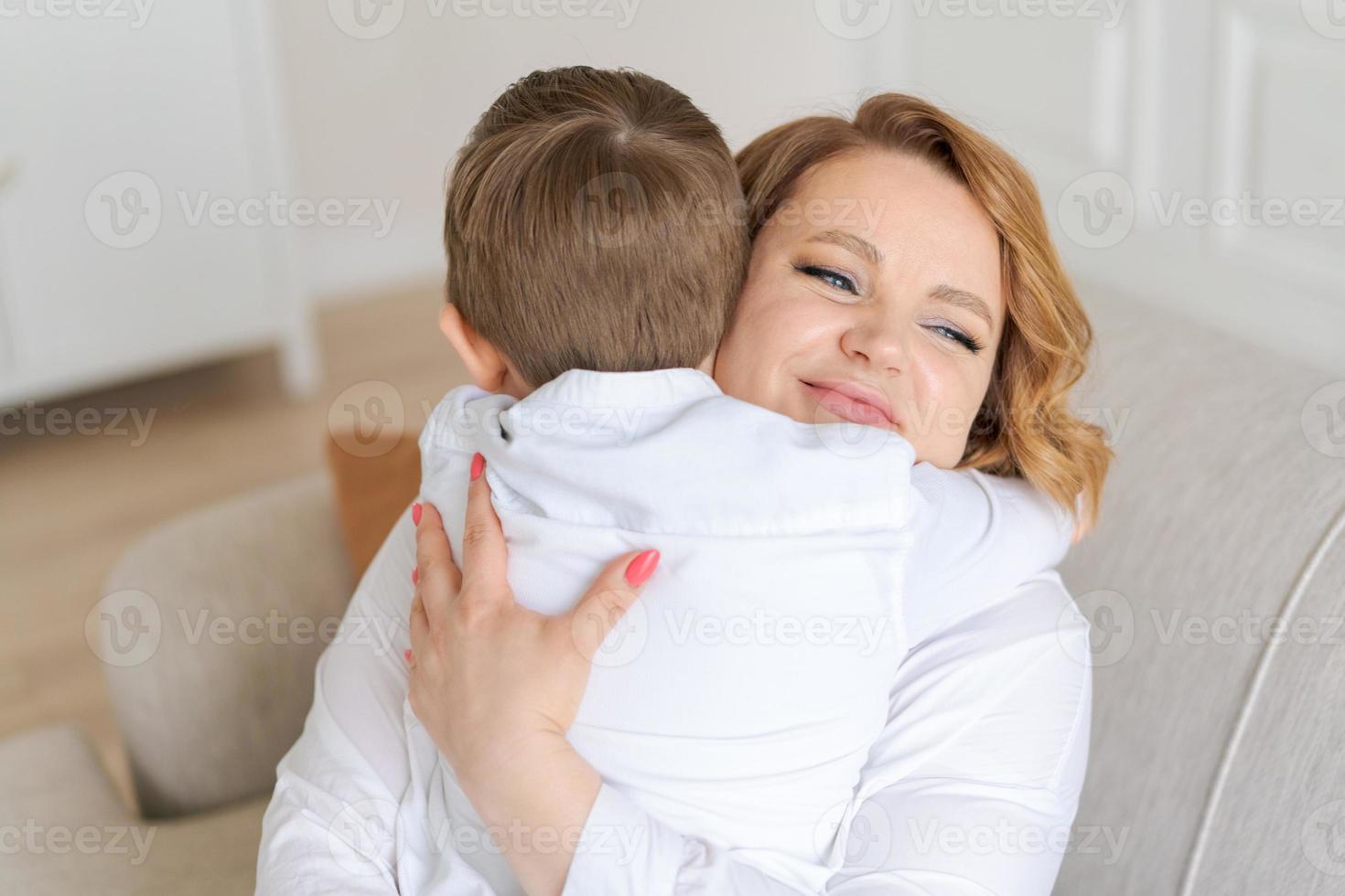 feliz filho caucasiano fofo abraça a mãe enquanto está sentado no sofá na sala de estar foto