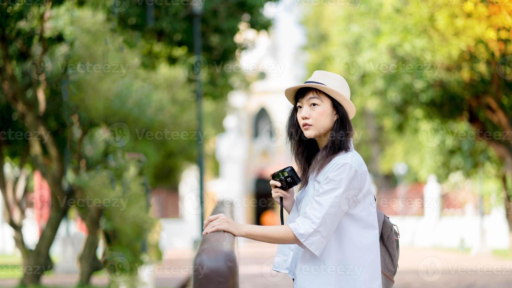 jovem viajante asiática com mochila veste camisa branca e chapéu segurando a câmera fotográfica na ponte foto