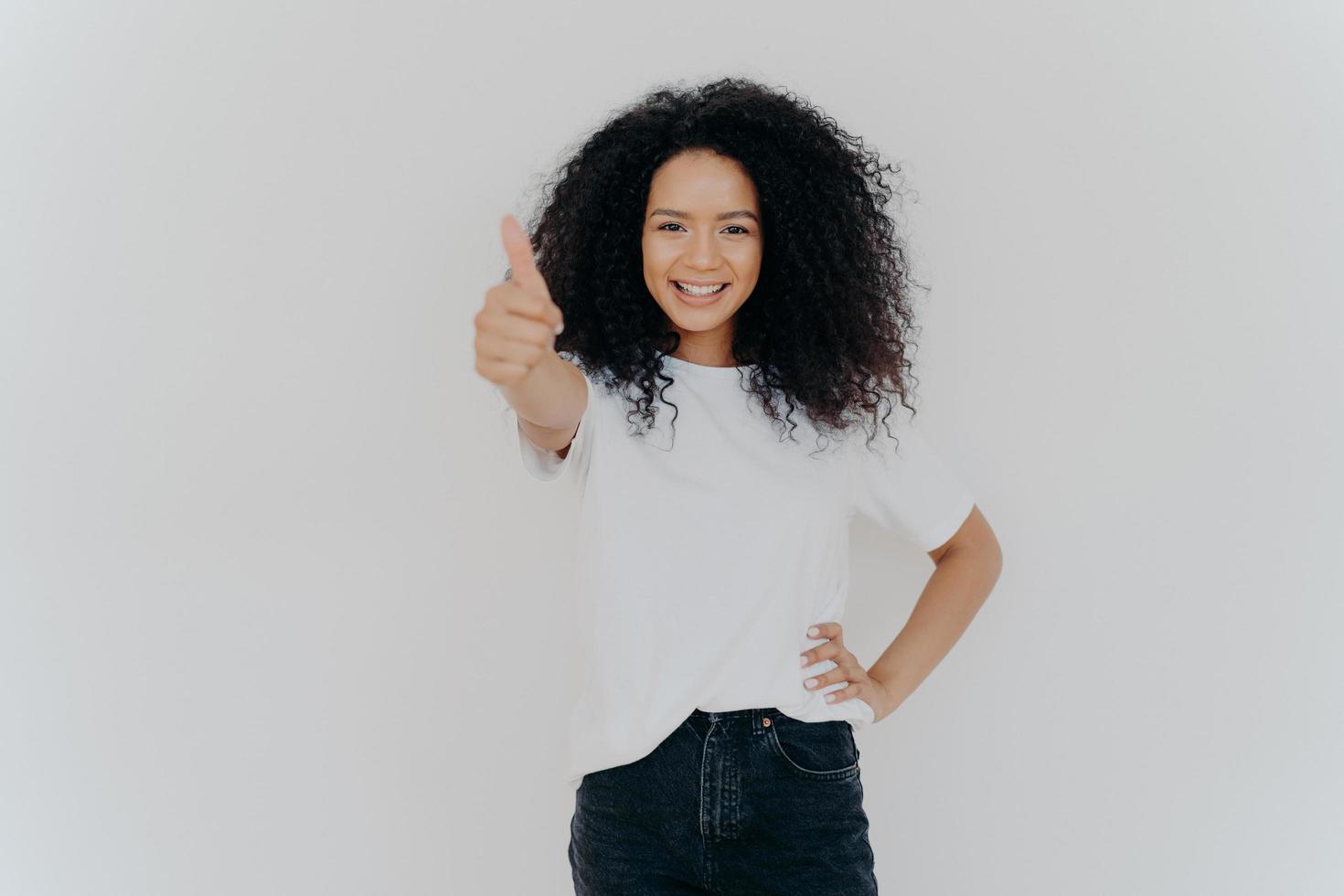 foto de mulher encaracolada feliz com cabelo crespo levanta o polegar, dá aprovação, diz que soa bem, faz gesto de apoio, mantém a outra mão na cintura, vestida casualmente, isolada sobre fundo branco