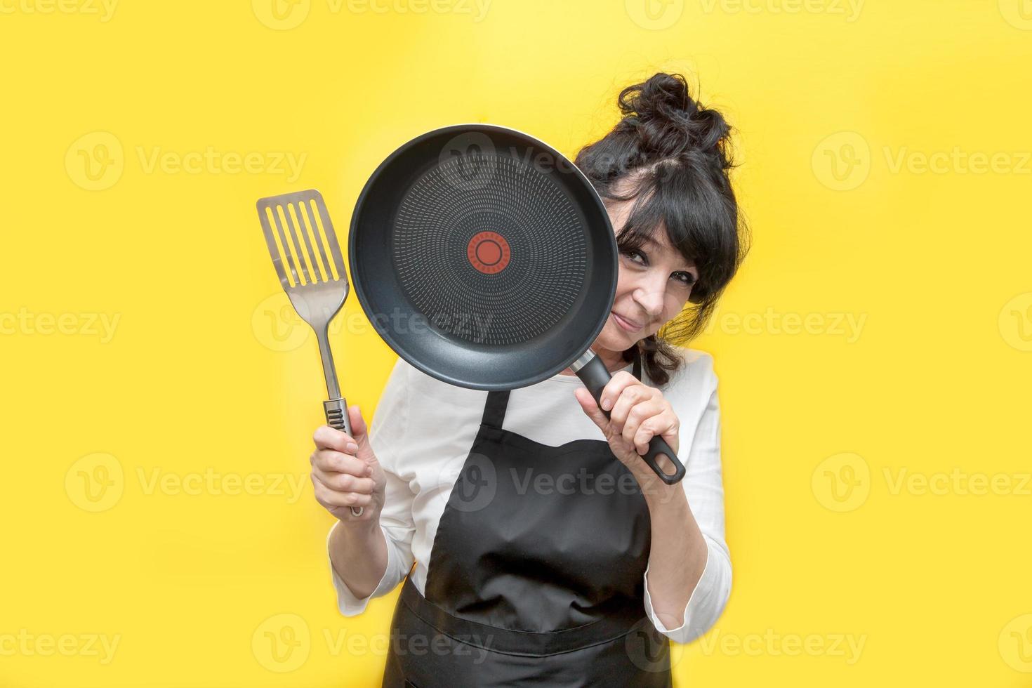 cozinheira idosa espreita por trás de uma frigideira que ela segura na mão, na segunda mão uma espátula foto