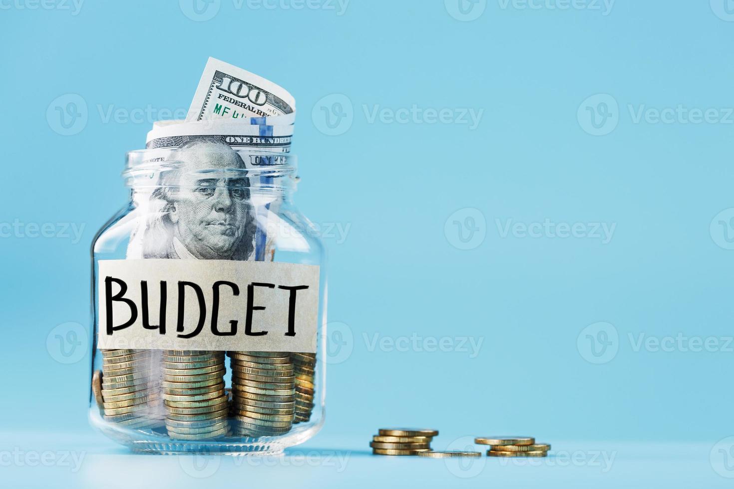 frasco de vidro com moedas e dólares, com um adesivo dizendo orçamento, sobre fundo azul. foto