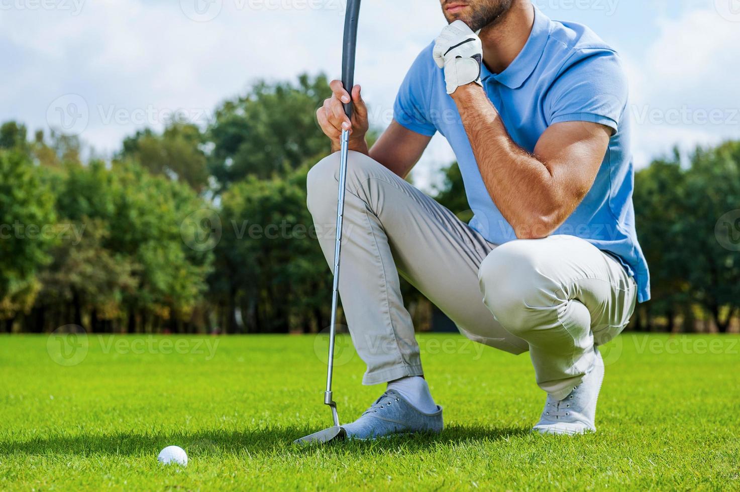 pensando no próximo tiro. imagem recortada de jovem jogador de golfe ajoelhado perto da bola de golfe e segurando a mão no queixo foto