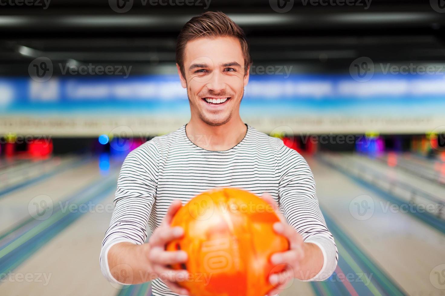 confiante e criativo. jovem alegre esticando uma bola de boliche e sorrindo em pé contra pistas de boliche foto