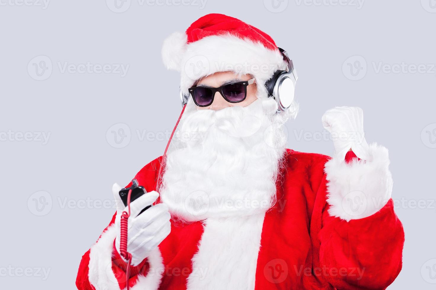 curtindo a música de natal. papai noel em óculos de sol e fones de ouvido ouvindo mp3 player e gesticulando em pé contra um fundo cinza foto