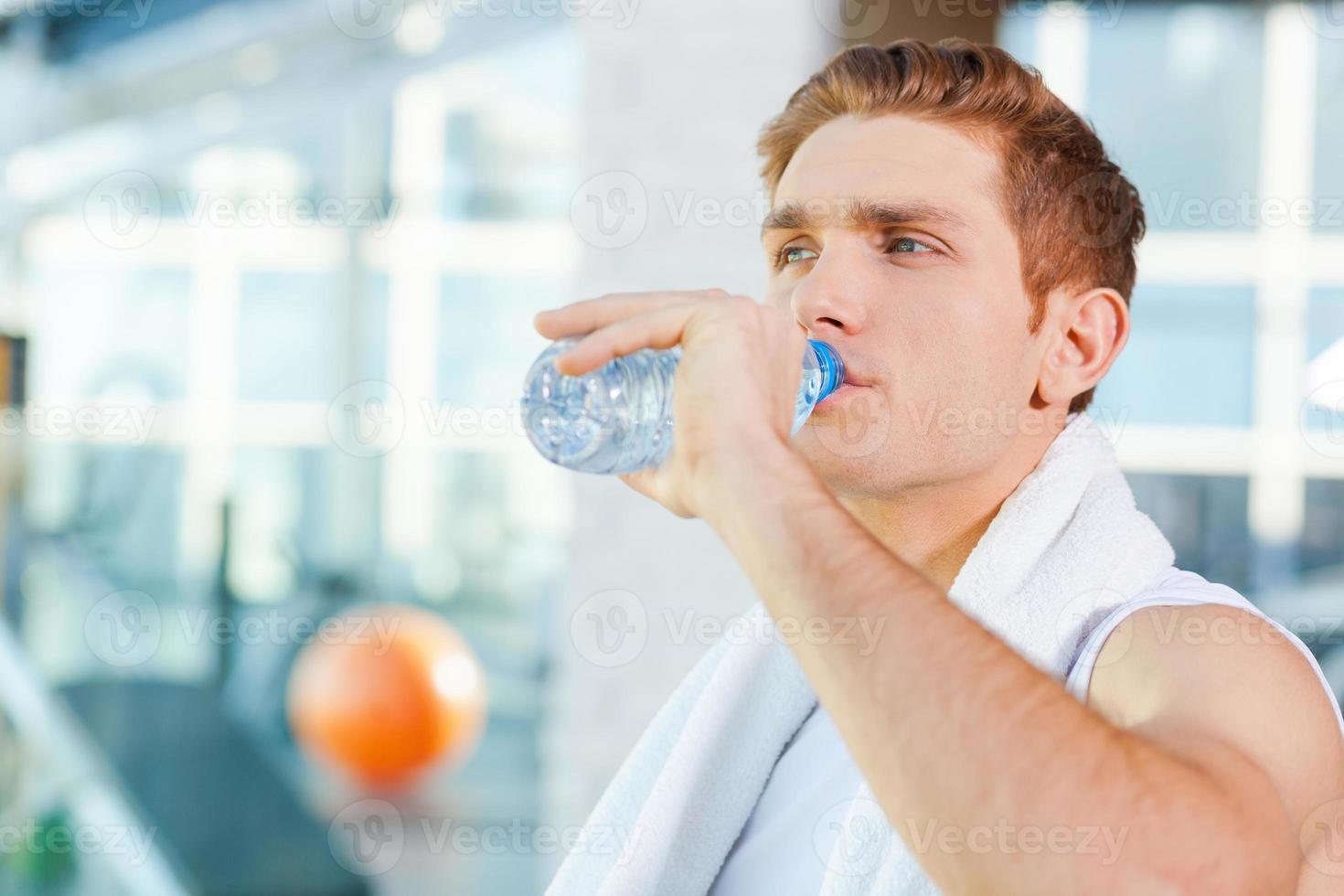 refrescante após o treino. jovem cansado carregando toalha nos ombros e água potável em pé no ginásio foto