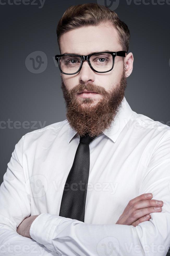 trazendo alguma criatividade para os negócios. bonito jovem barbudo de camisa e gravata, mantendo os braços cruzados e olhando para a câmera em pé contra um fundo cinza foto