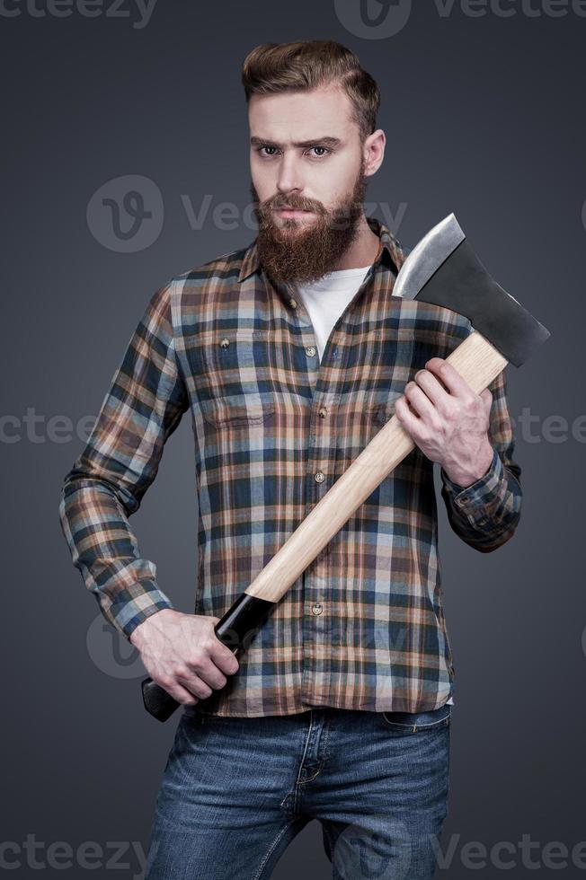 estilo lenhador. bonito jovem barbudo segurando um machado grande e olhando para a câmera em pé contra um fundo cinza foto