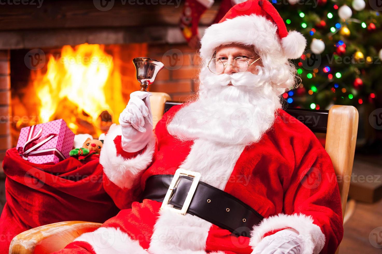 feliz natal e feliz ano novo alegre papai noel sentado em sua cadeira e tocando uma campainha com lareira e árvore de natal ao fundo foto