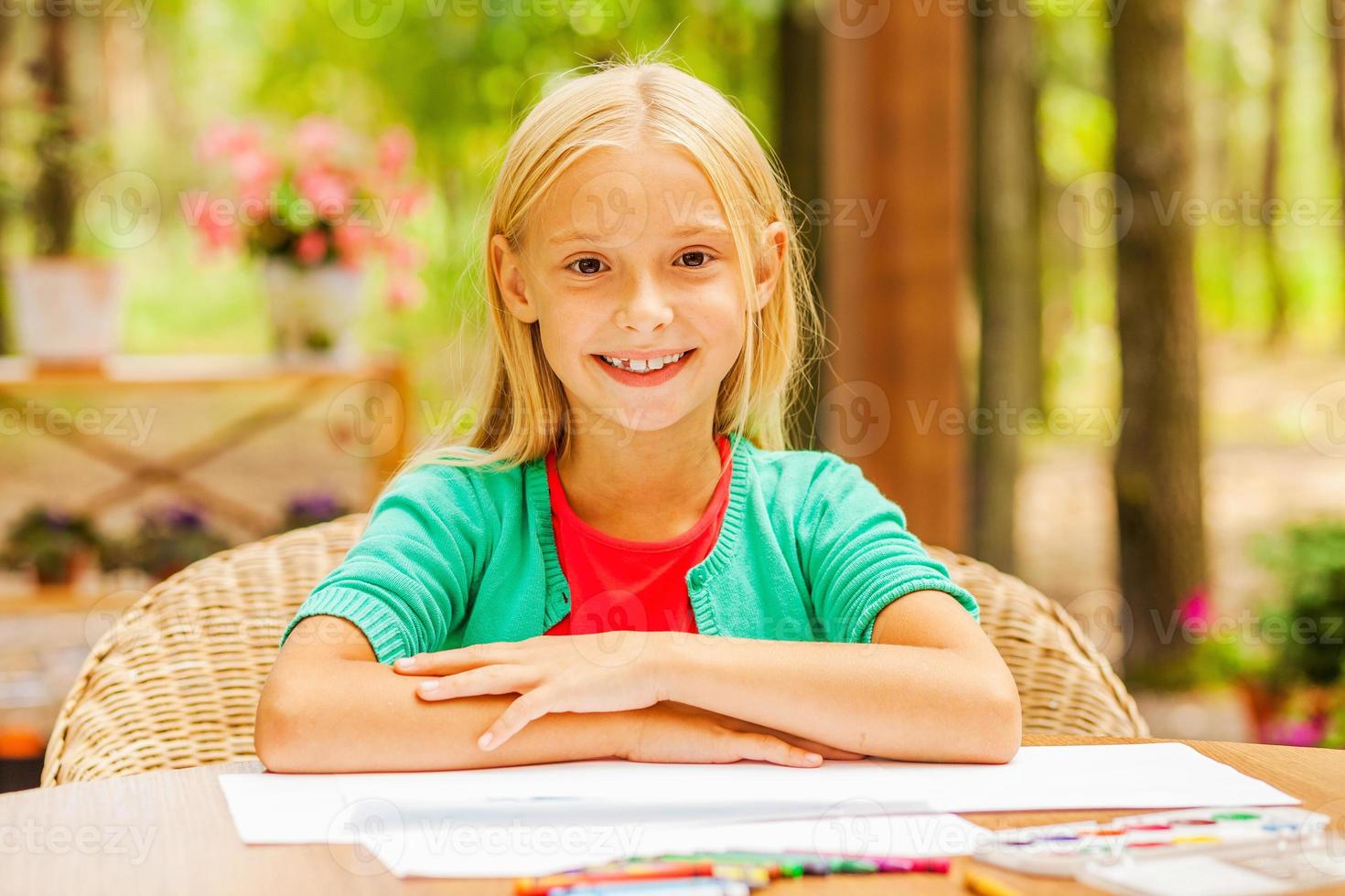 pequeno raio de sol. menina bonitinha olhando para a câmera e sorrindo enquanto está sentado à mesa com lápis coloridos e papel deitado sobre ele foto