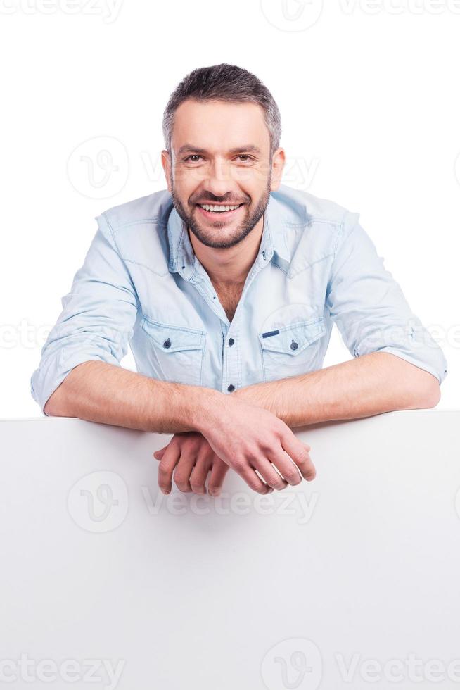 sua publicidade abaixo. jovem bonito em roupas casuais, inclinando-se no espaço da cópia e sorrindo em pé contra um fundo branco foto