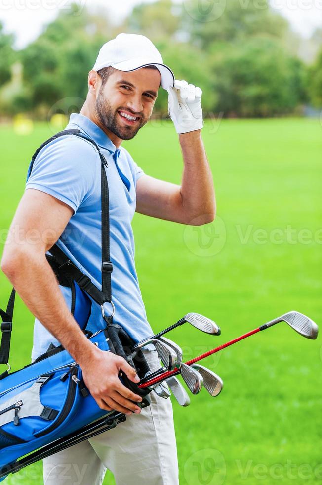 eu amo golfe vista traseira do jovem golfista feliz carregando saco de golfe com motoristas e olhando por cima do ombro em pé no campo de golfe foto