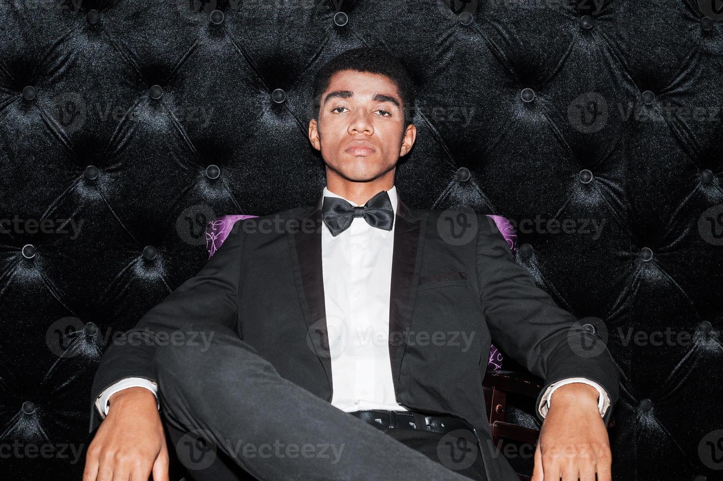 confiante de ser o melhor. elegante jovem afro-americano sentado na cadeira colocando uma perna na outra foto