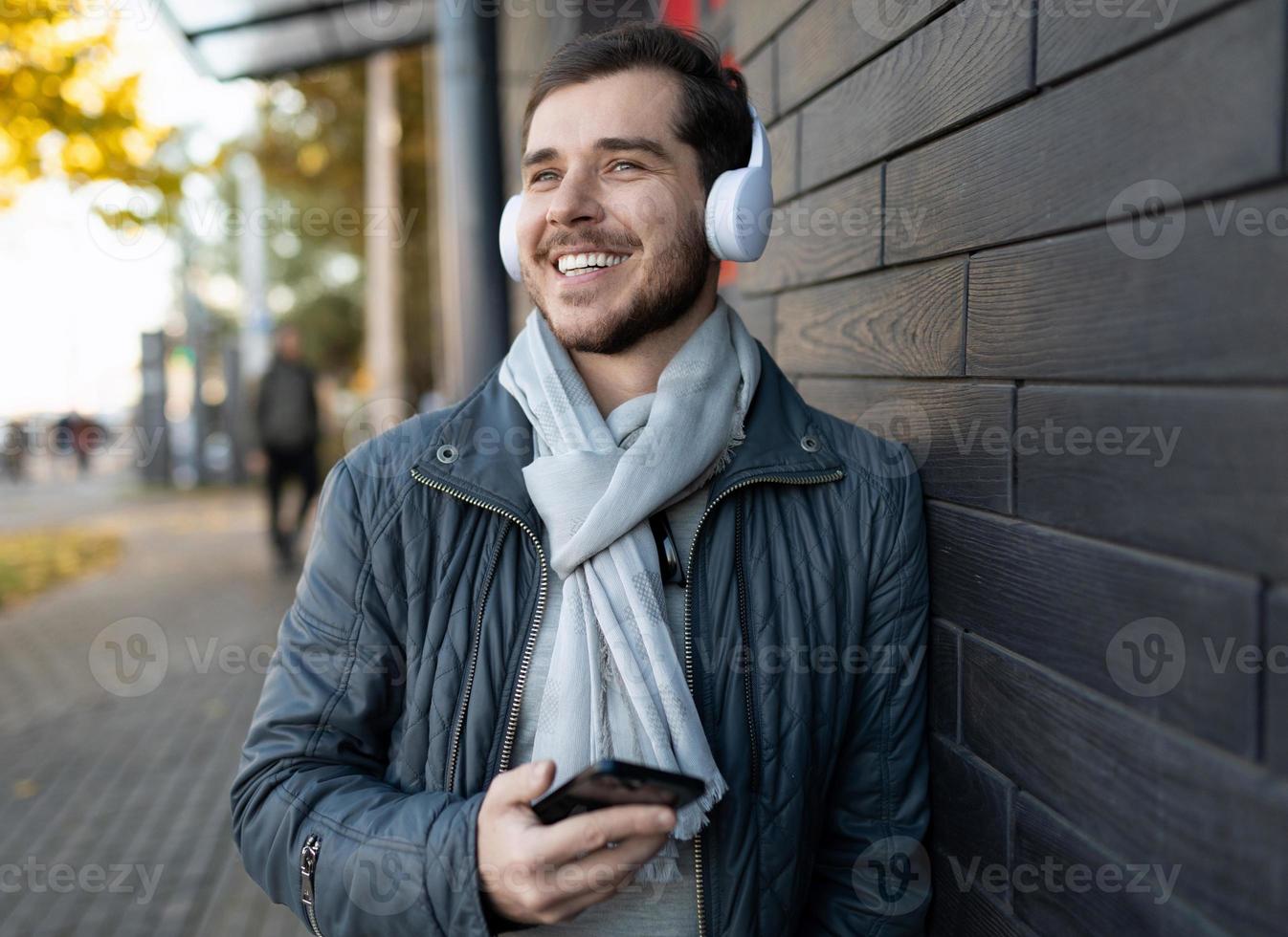 um homem adulto alegre ouve música em fones de ouvido brancos em um telefone celular e olha para longe com um sorriso foto